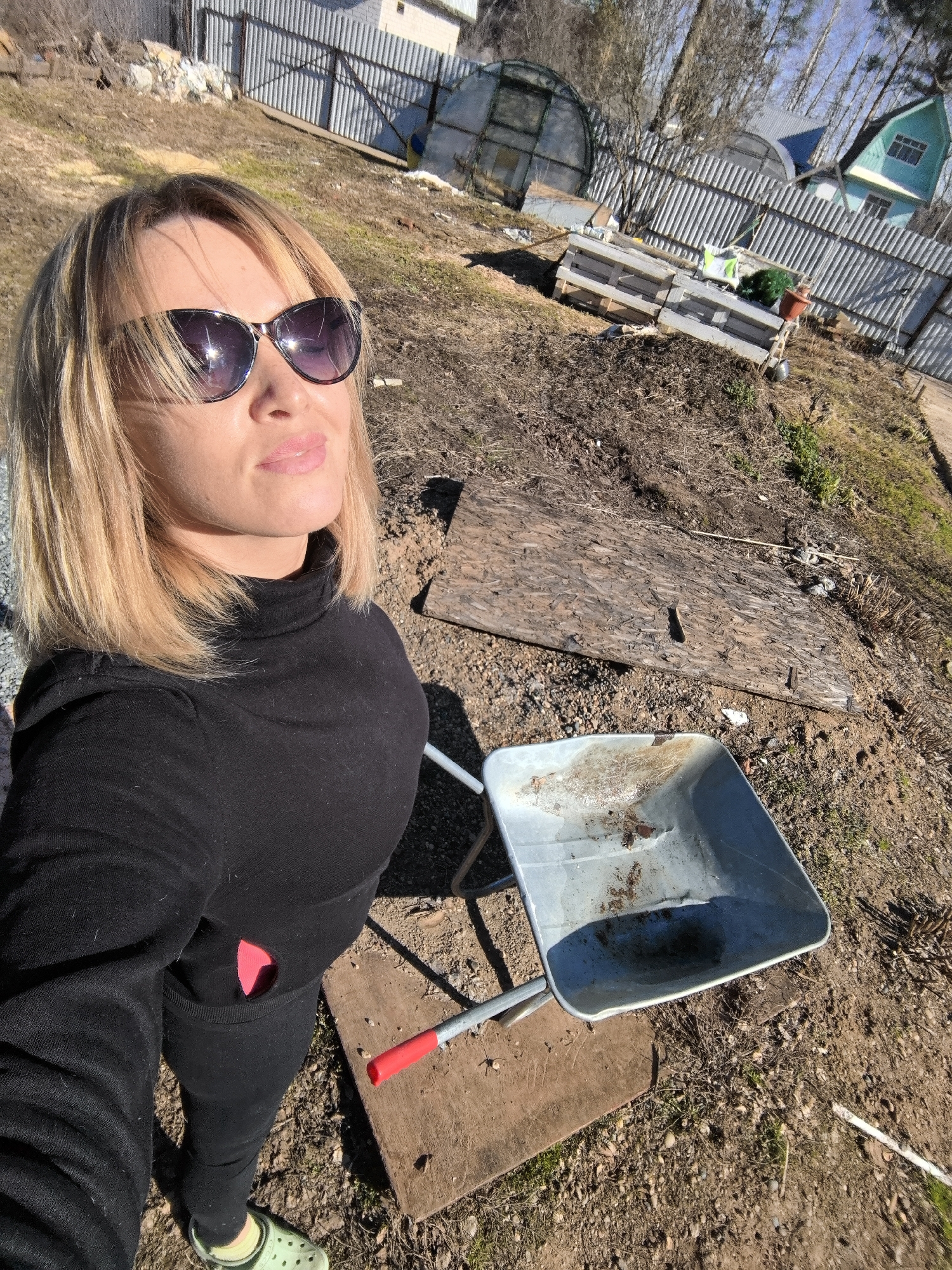 A blonde woman, wearing sunglasses, a black turtleneck, black pants with a pink patch, and light green Crocs, is the central subject of this outdoor selfie. Her head is tilted slightly upwards, and her expression appears relaxed under the bright sunlight.
The setting is a rustic outdoor area in Noviy, Russia, likely a garden or yard. The ground is covered in dry dirt and sparse, dry grass, characteristic of early spring. An empty, well-used metal wheelbarrow with a red handle is positioned near the woman, resting on a piece of wood. Several other wooden planks are scattered on the ground.
In the background, a corrugated metal fence separates the foreground from distant structures. Beyond the fence, two arched greenhouse-like structures are visible, along with a light blue or green house with a distinctive roofline. Stacks of wooden pallets and a pot holding a small green plant are also seen near the fence. The clear sky and bright sun suggest it is a sunny day. No text is visible in the image. The scene suggests a casual moment during outdoor work or leisure in a rural or suburban environment.