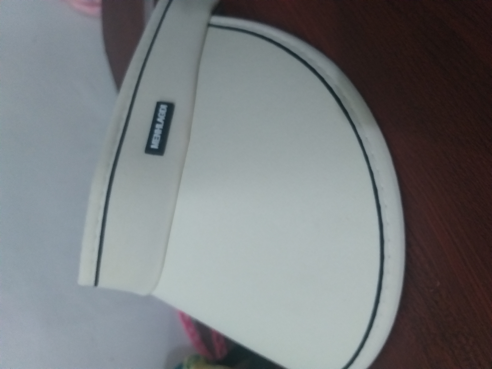 The image shows a close-up of a white visor with a black trim and a small black label with white lettering that reads "MENHLAGDI". The visor is placed on a brown wooden surface. The background is blurred and appears to be a light-colored wall, with some colorful objects visible on the left side, possibly fabric. The lighting is soft, suggesting an indoor setting. There are no people visible in the image. The location in Air Jamban, Indonesia is not discernible from the visual information provided.