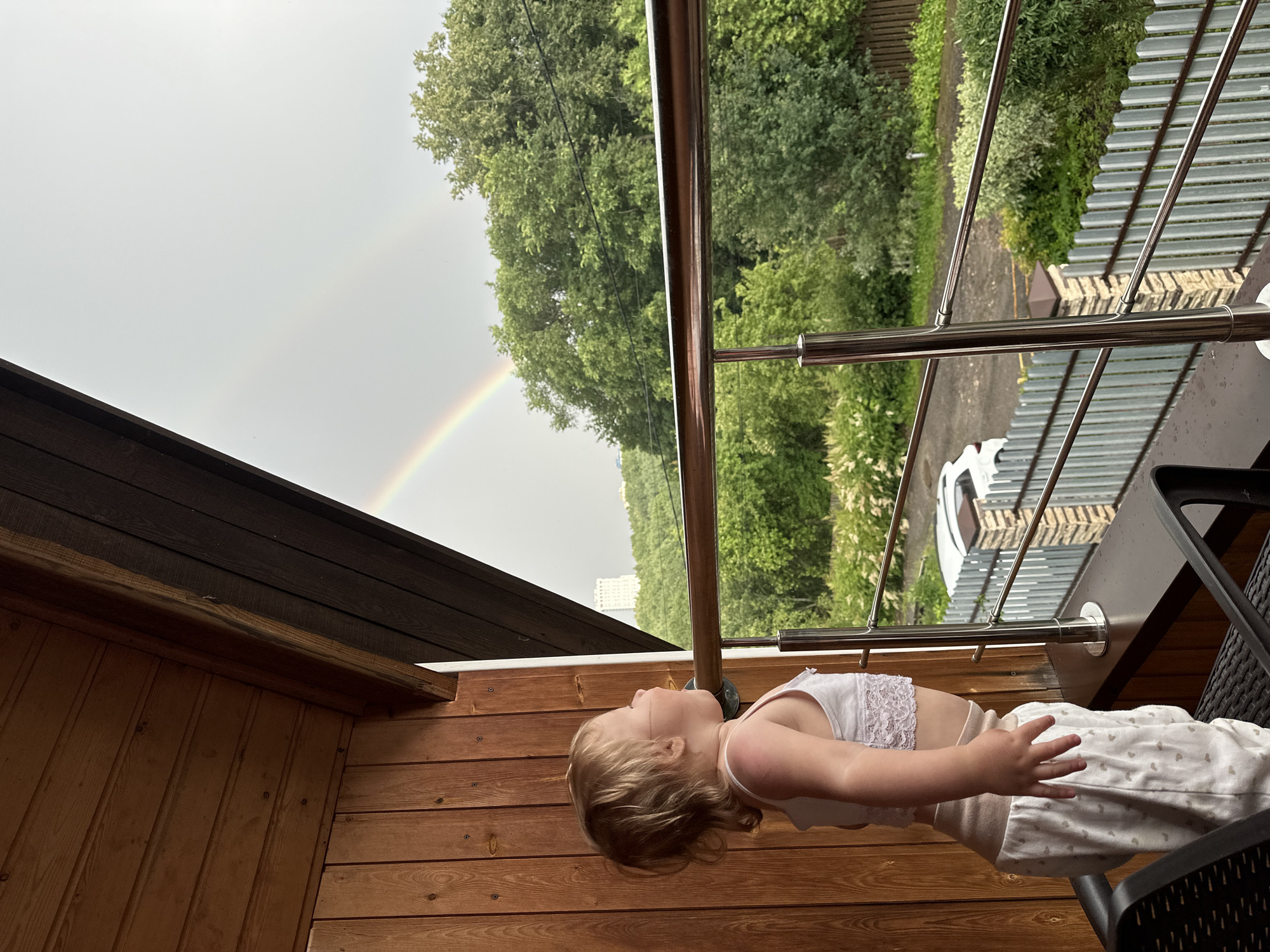 A young child with blonde hair is lying on a wooden balcony, looking up at a rainbow in the sky. The child is wearing a white lace-trimmed top and patterned pants. The balcony has a wooden railing and a dark wooden roof overhang. Beyond the balcony, lush green trees and a corrugated metal fence are visible. The sky is overcast, with the rainbow prominently displayed. The overall impression is one of peaceful observation of nature after a rain shower. The image appears to be taken during daylight hours, with the soft light suggesting it could be late afternoon or early morning.