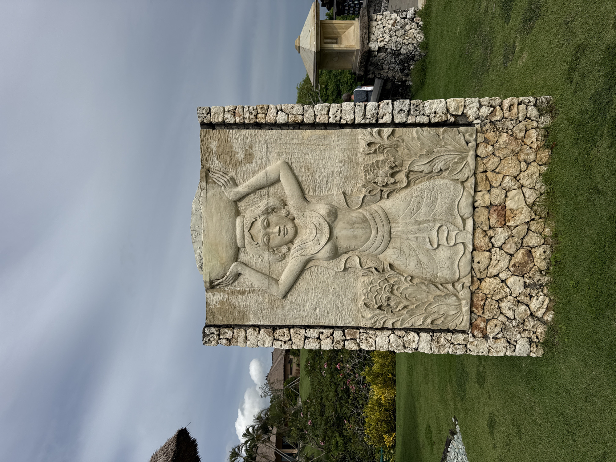 The image shows a stone carving of a woman. The carving is surrounded by a border of rough-hewn stones. The background includes green grass, trees, and a small building. The sky is overcast. This appears to be located in Kuta Selatan, Indonesia.