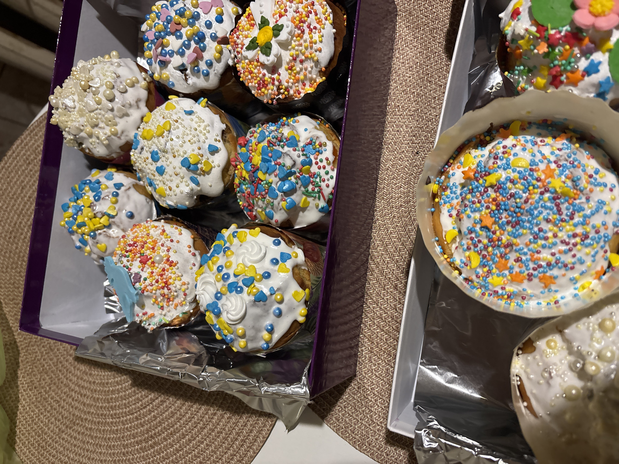 Two open boxes of frosted and decorated Easter cakes sit on a table covered with a textured, tan-colored surface. The box on the left is purple and contains ten cakes, each with different colored sprinkles and decorations on a white frosting base. The box on the right is white and holds two cakes, also decorated with sprinkles, one with a large variety of colors and another primarily with pearl-like sprinkles. The cakes are arranged on aluminum foil within the boxes. The location is Pokotylivka, Ukraine.