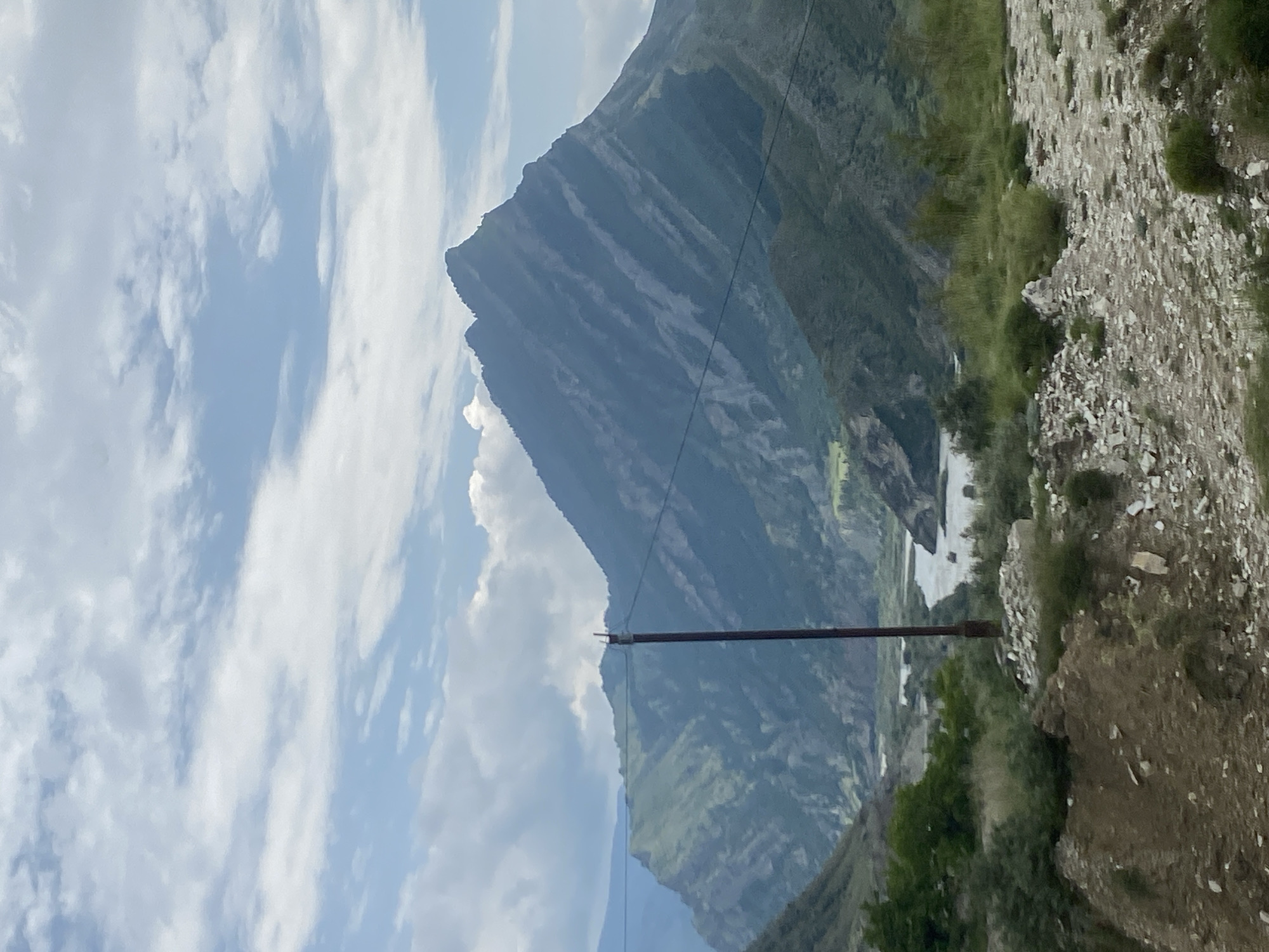 The image depicts a mountainous landscape with a river winding through a valley under a cloudy sky. In the foreground, a dirt path with scattered rocks and sparse vegetation leads towards the middle ground. A single, tall, dark pole with wires stretches across the scene, partially obscuring the view of the mountains and river. The mountains themselves are rugged and covered in green vegetation, with some rocky outcrops visible. The river appears to be flowing through a gorge, with lighter-colored water. The overall impression is of a natural, somewhat remote, and undeveloped area. The metadata indicates the location as Chalda, Russia.