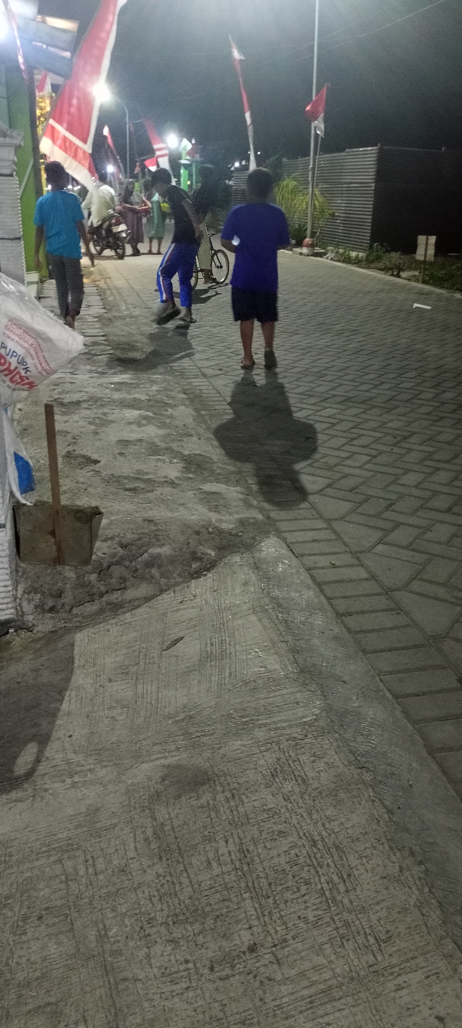 In Gempolklutuk, Indonesia, a group of people are gathered outdoors at night. The scene appears to be on a paved walkway with a concrete area adjacent to it. Several Indonesian flags are visible, suggesting a possible celebration or national event.
In the foreground, to the left, a large white sack with text printed on it is partially visible. Next to it, a shovel with a wooden handle rests in a shallow indentation in the concrete. The concrete surface displays a rough, textured finish.
Further into the image, three young males are prominently featured. One, wearing a blue t-shirt and grey pants, walks away from the camera. Another, in a dark t-shirt and blue pants with white stripes, is in motion, possibly kicking or playing. A third individual, in a blue t-shirt and dark shorts, stands facing away from the viewer.
Behind these individuals, a motorcycle is parked, and other figures, including adults, are gathered. The background shows a dark, possibly wooded area, and a corrugated metal fence. The lighting suggests artificial light sources, illuminating the area and casting shadows on the ground.