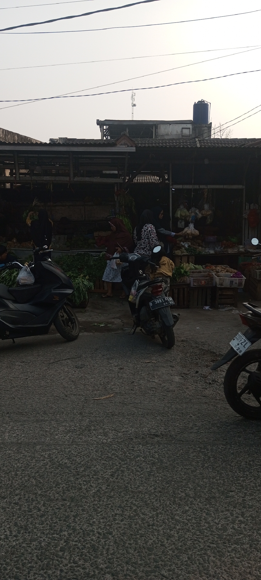 The image depicts an outdoor scene at a market, likely a roadside market given the presence of motorcycles and a paved road. The market stalls, constructed with wood and corrugated metal roofing, display a variety of produce, including what appear to be root vegetables and leafy greens. Several individuals are present, engaged in shopping or attending to the stalls. One woman in a hijab is handing items to another, while a child in a yellow top is visible. Two motorcycles are parked in the foreground, one dark-colored and the other grey, with license plates indicating they are registered in Indonesia. The background shows a building with a blue water tank and a telecommunications tower, suggesting an urban or semi-urban environment. The sky is overcast with a hint of haze. The overall impression is of a busy, functional marketplace in Setu, Indonesia.