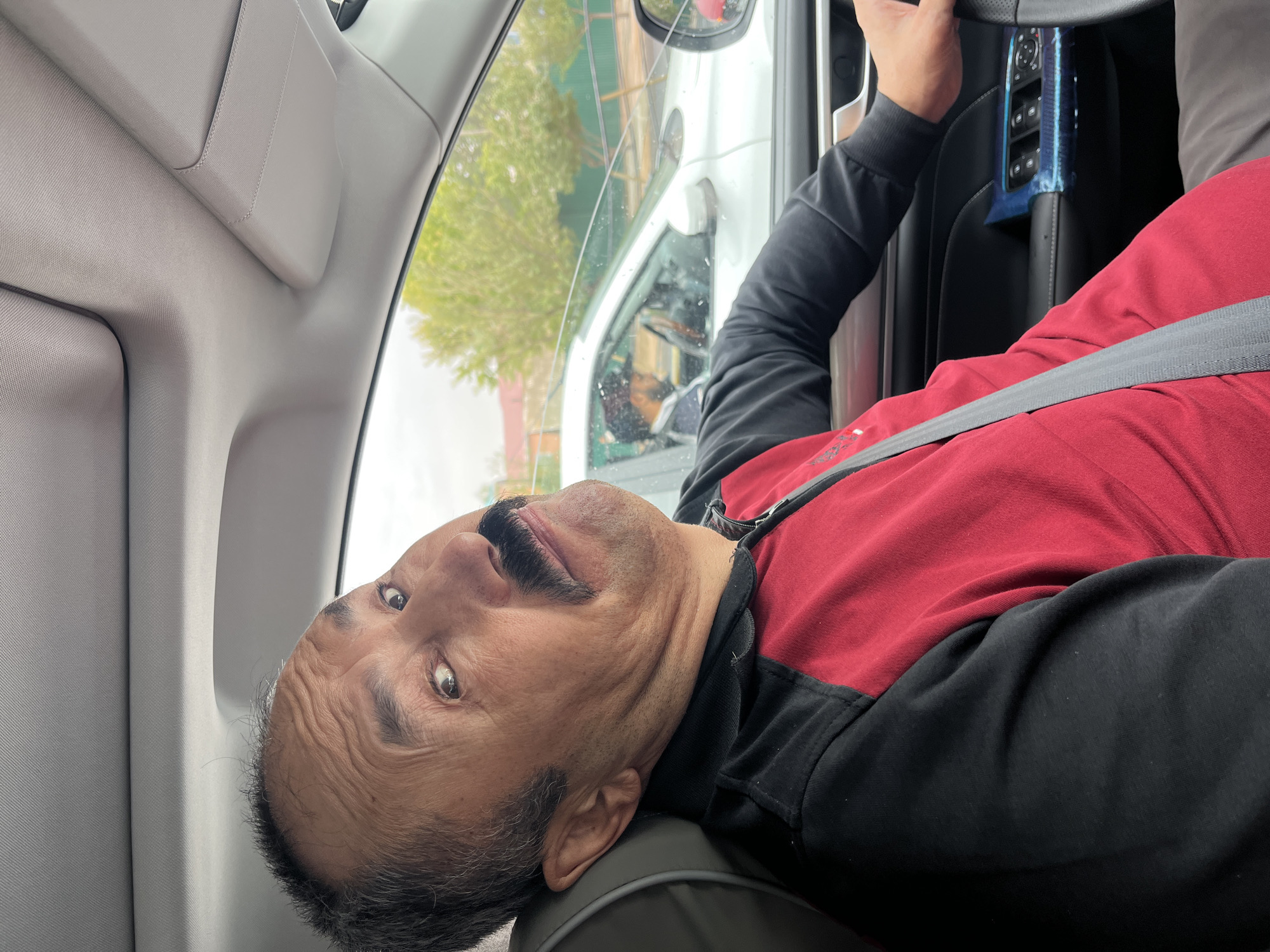 A man with a mustache is driving a car. He is looking at the camera with a surprised expression. He is wearing a red and black jacket. The car is in motion, and another car is visible in the background. The scene appears to be in Astana, Kazakhstan, as indicated by the architecture and vegetation. The car's interior is modern, with leather seats and electronic controls. The lighting suggests it is daytime.
