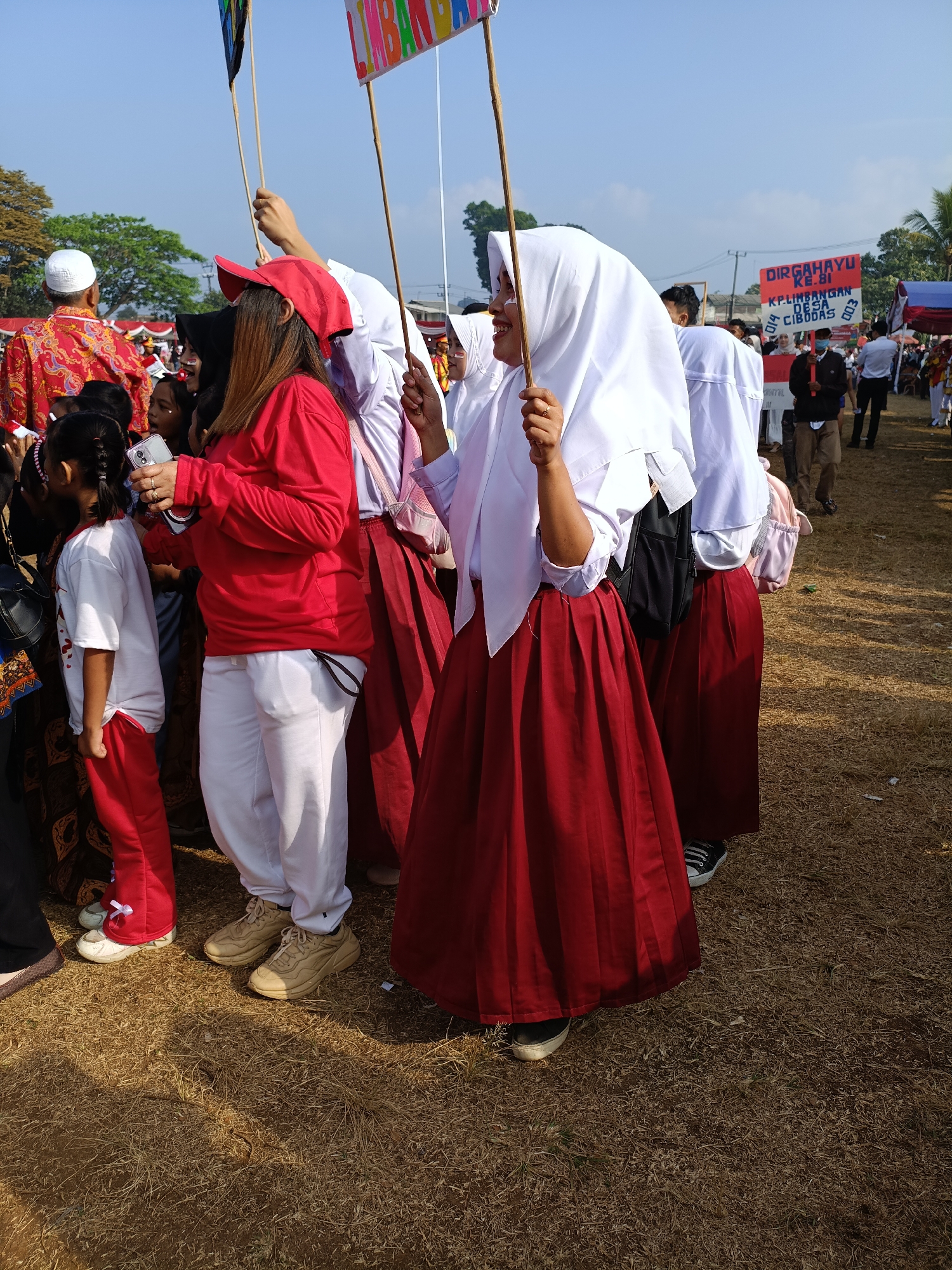 In Cibodas, Indonesia, a group of students in uniform stand on dry grass, participating in an outdoor event. Several girls, wearing white hijabs and red skirts, are visible. One girl in the foreground holds a wooden stick with a banner that says "LIMBANG". Another girl, also in a white hijab and red skirt, is to her right. A woman in a red shirt and white pants, wearing a red cap, is to the left, holding a mobile phone. Behind them, other people are gathered, with a banner that reads "DIRGAHAYU KE-BI". The scene suggests a celebratory occasion, possibly a parade or school event, under a clear blue sky.