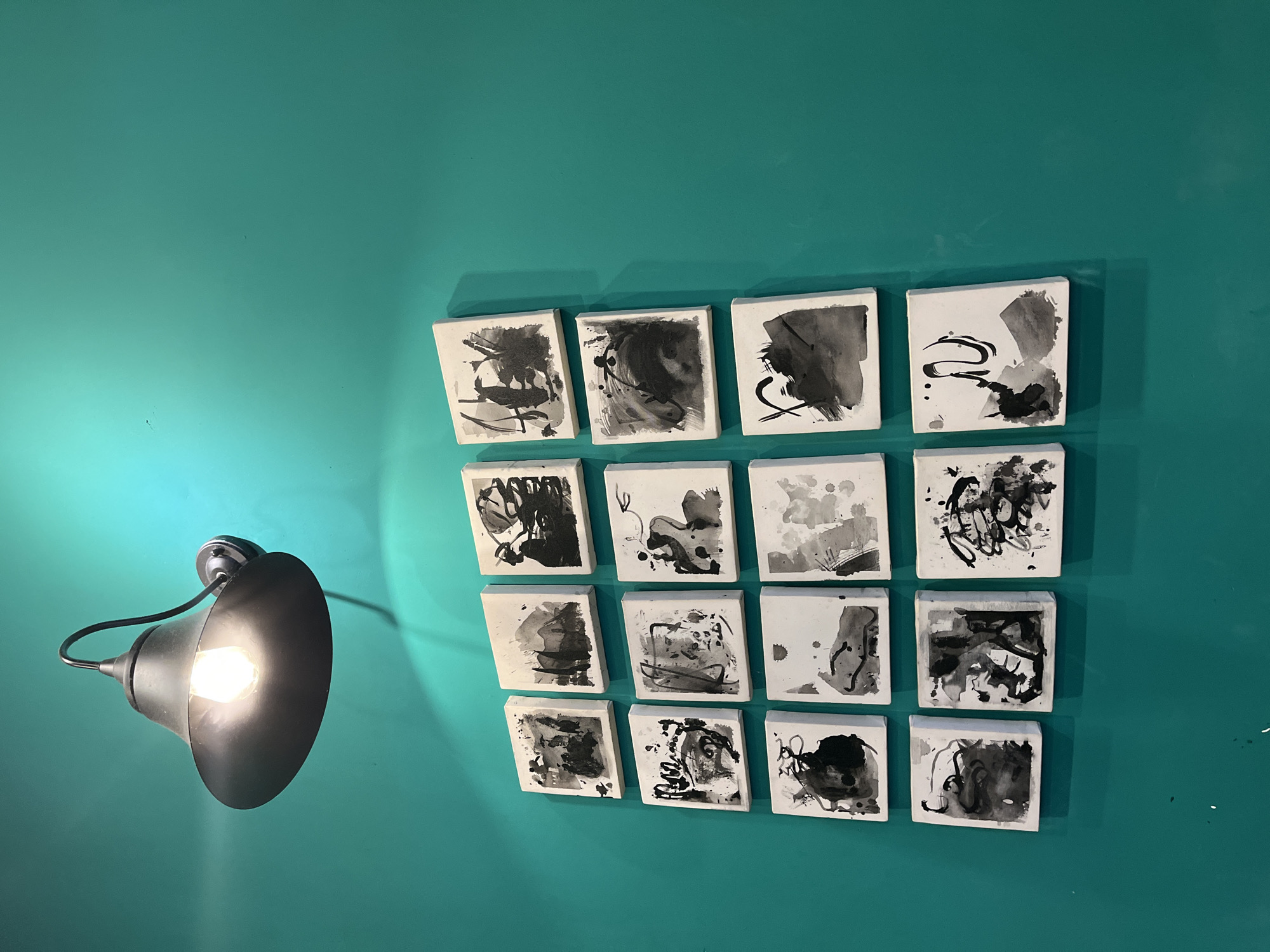 The image displays an interior wall painted in a vibrant teal or turquoise color, featuring a modern wall lamp and a grid of abstract art pieces. On the left side, a dark-colored, industrial-style wall lamp extends from the wall with a curved arm, holding a visible, brightly glowing incandescent bulb. The lamp casts a warm, focused light, creating a bright spot on the wall that subtly fades into the ambient teal.
To the right, a symmetrical arrangement of 16 small, square art pieces is mounted on the wall, forming a 4x4 grid. Each piece features unique abstract designs executed in black and shades of gray on a white background, reminiscent of ink wash paintings or abstract expressionism. The artworks show spontaneous brushstrokes, splatters, and fluid forms, creating a visually dynamic collection. Shadows from these art pieces are visible on the wall, cast by the light from the lamp.
There are no people or specific activities depicted. No text is visible within the frame. The setting appears to be an indoor space, possibly a contemporary home, gallery, or cafe in Đà Nẵng, Vietnam, where the vibrant wall color and art selection contribute to a distinct aesthetic. The illumination from the lamp suggests it might be evening or a space where artificial lighting is primarily used.