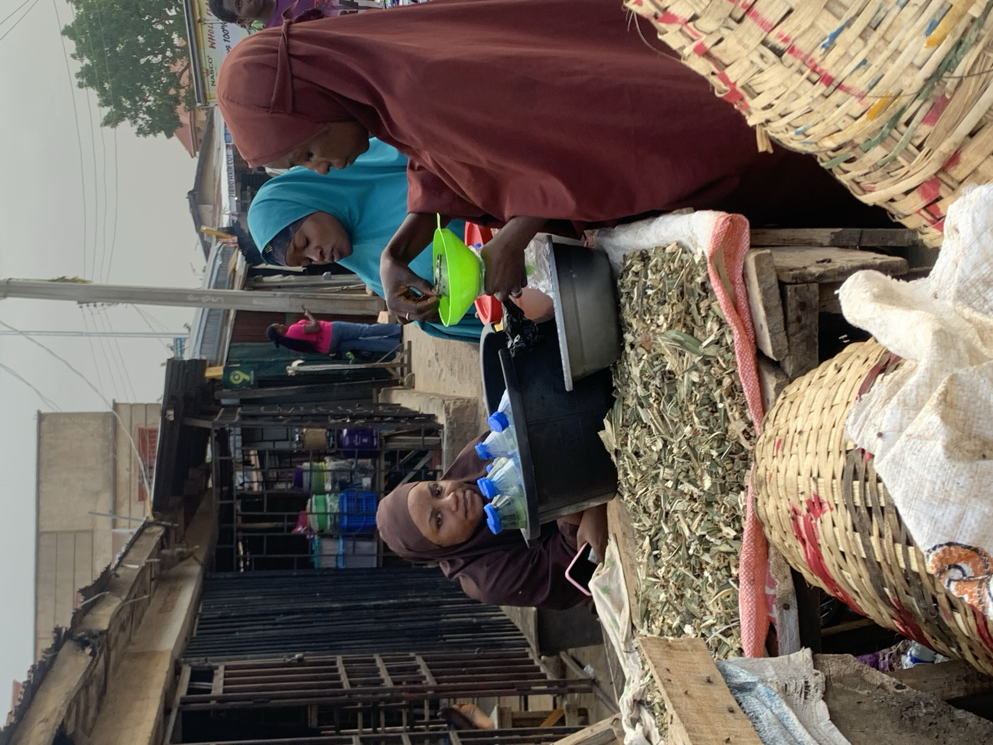 In a street-level market in Jos, Nigeria, two women in headscarves, one brown and one blue, are engaged in a transaction involving dried leaves. The blue-scarved woman uses a green funnel to fill small plastic bottles from a black container, as the brown-scarved woman watches. A black pail filled with plastic bottles stands nearby. Dried leaves are displayed alongside, with woven baskets visible in the foreground. Behind them, market stalls and urban structures are observable in the background.