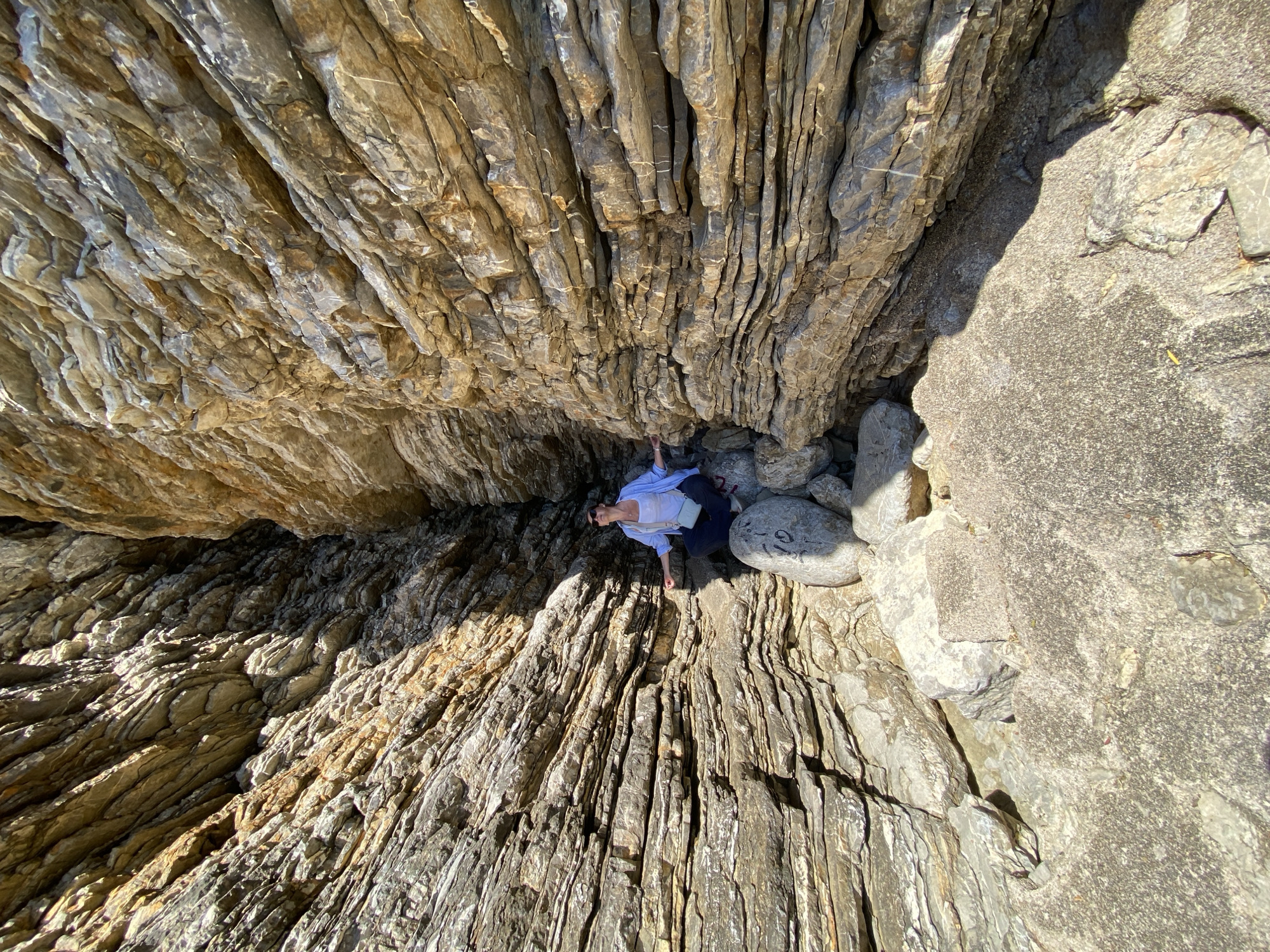 An overhead photograph captures a person nestled within a deep, narrow rock formation in Budva, Montenegro. The scene is dominated by towering, layered rock walls that rise on either side, creating a cave-like ambiance. These rocks display pronounced stratification and texture, with a palette of greys, browns, and warm ochre tones, indicating complex geological features. Intense, direct sunlight illuminates the rock surfaces, casting sharp shadows and highlighting their intricate details, suggesting a bright, clear day.
In the lower center of the frame, a person, appearing to be a woman, is seen sitting or crouching amongst scattered boulders, looking upwards. She is dressed in a light blue long-sleeved shirt over a white top and dark trousers. Her right hand rests on a smooth, lighter-colored rock. On a larger, rounded boulder positioned to her right, faint, dark markings are discernible, possibly numbers such as "19". The unique perspective emphasizes the scale and rugged beauty of the natural rock structure, with the individual providing a sense of depth and human interaction within the formidable landscape.