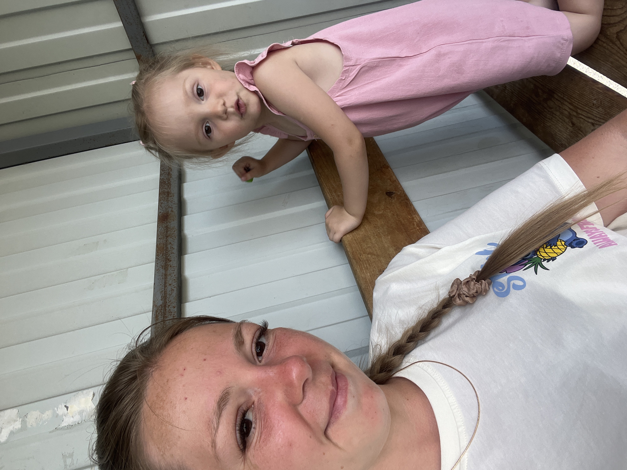 This is a selfie taken outdoors, likely in a rural or semi-rural setting in Shadrinsk, Russia. The image features two individuals: a young child and an adult. The child, a girl with blonde hair and fair skin, is wearing a pink sleeveless dress. She is positioned higher in the frame, looking towards the camera with an open-mouthed expression. She appears to be leaning on a wooden plank. The adult, whose face is closer to the camera and takes up a significant portion of the frame, has brown hair styled in a braid and is wearing a white t-shirt with a graphic print. Her expression is relaxed and she seems to be smiling or smirking.
The background consists of corrugated metal siding, suggesting a structure like a shed or outbuilding. A metal support beam is also visible. The lighting suggests it is daytime, and the overall atmosphere appears casual and informal, possibly indicating a family outing or a moment of play. The adult's t-shirt has visible text and graphics, including what appears to be a pineapple and possibly a cartoon character, although the text is not clearly legible. The image is taken from a low angle, looking up at the child and then towards the adult.