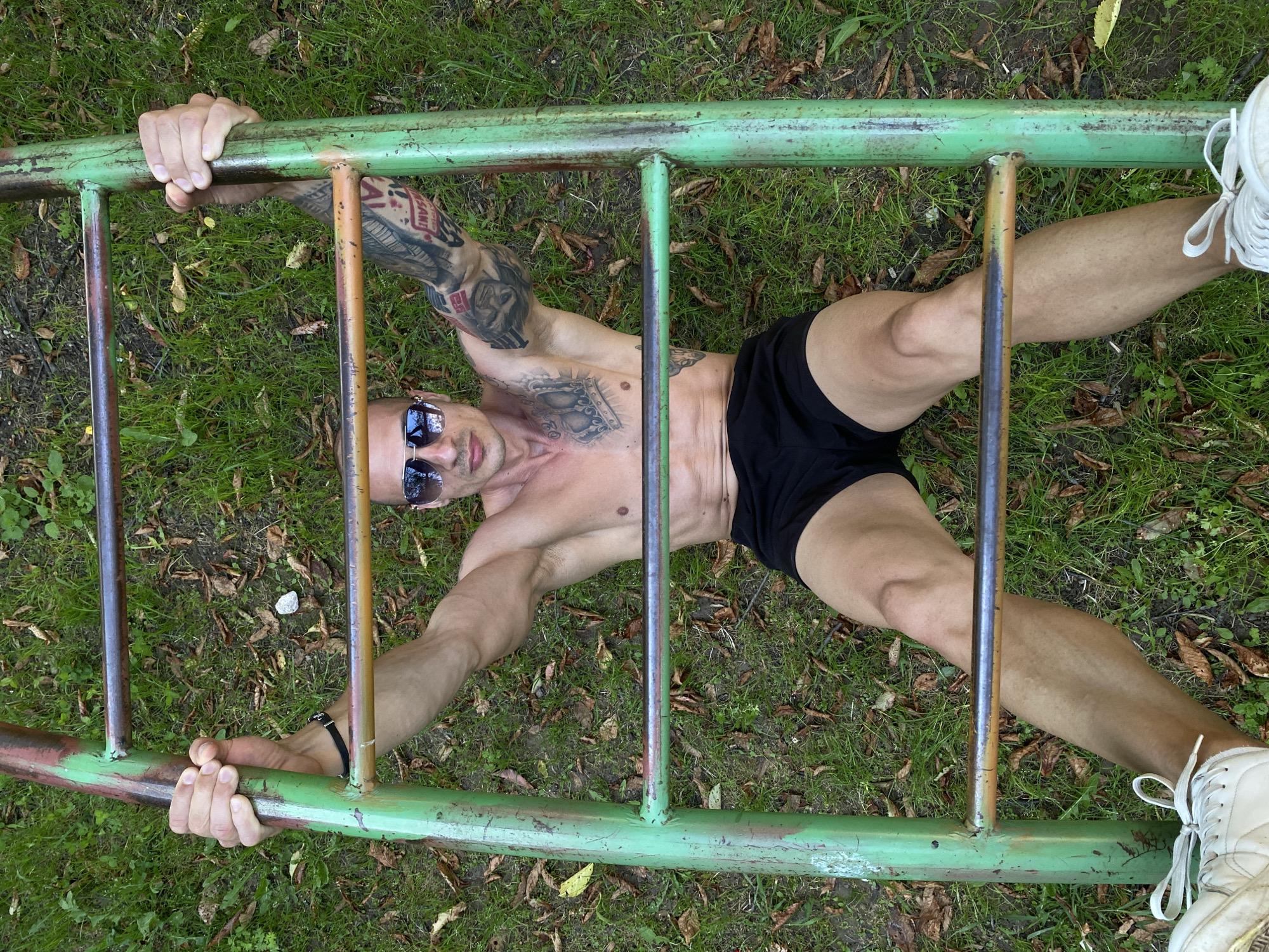 A high-angle shot depicts a muscular adult male lying on his back on a grassy surface, looking directly upwards at the camera. He is positioned beneath an aged, green and rust-colored metal ladder-like structure, gripping one of its horizontal rungs with both hands, his arms extended.
The man is shirtless, revealing a crown tattoo on his upper right chest and an extensive sleeve tattoo on his left arm that includes the text "SANT". He has a bald or closely shaven head and wears reflective aviator-style sunglasses. A black bracelet is visible on his left wrist. He is dressed in black athletic shorts and white sneakers with white laces.
The ground beneath him is covered with green grass, significantly interspersed with dry brown and yellow fallen leaves. A small white rock rests on the grass near the top center of the frame. The metal structure consists of two vertical supports and multiple horizontal rungs, showing considerable wear, chipped paint, and visible rust, indicating its age and outdoor exposure. The scene is an outdoor public green space, likely a park or an area with recreational equipment. This outdoor scene is situated in the city of Pleven, Bulgaria.