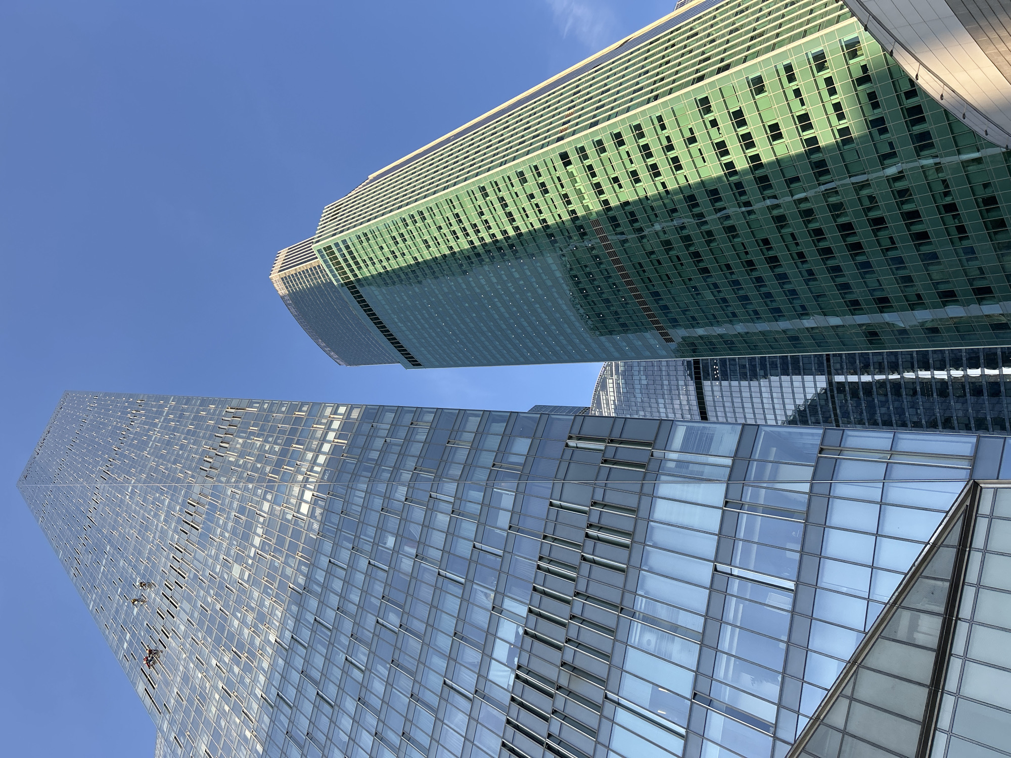 The image shows a low-angle view of two skyscrapers against a clear blue sky. The building on the left has a predominantly glass facade, reflecting the sky, and window washers appear to be working on the building. The building on the right has a greenish tint and a regular grid of windows, with a shadow diagonally covering a section. The location appears to be an urban area, with the composition focusing on the height and architecture of the buildings.