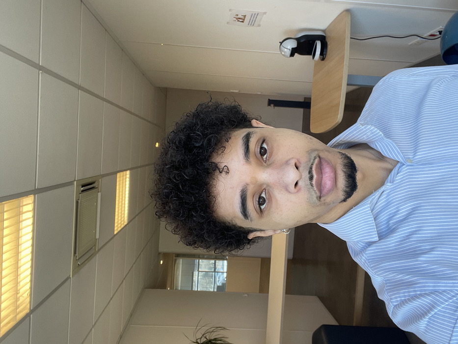 A light-skinned male, likely in his twenties, with dark curly hair, a mustache, and a goatee, is depicted in a self-portrait. He wears a light blue and white striped collared shirt and a silver hoop earring in his left ear. His brown eyes are directed toward the camera. The background is an interior office or commercial space with light-colored walls and a white grid-paneled ceiling containing recessed fluorescent light fixtures, some emitting a warm yellow light. An air conditioning vent is visible within the ceiling grid. To the subject's right, a black and white capsule coffee machine sits on a small, light wood wall-mounted table with a power cord descending along the wall. Above the coffee machine, a white paper notice with red text is affixed to the wall. To the subject's left, a light wood counter or desk structure is partially visible, along with a dark blue object in the lower corner. A doorway in the mid-background reveals a glimpse of an outdoor scene with vehicles and trees through a window. The location is Montigny-le-Bretonneux, France.