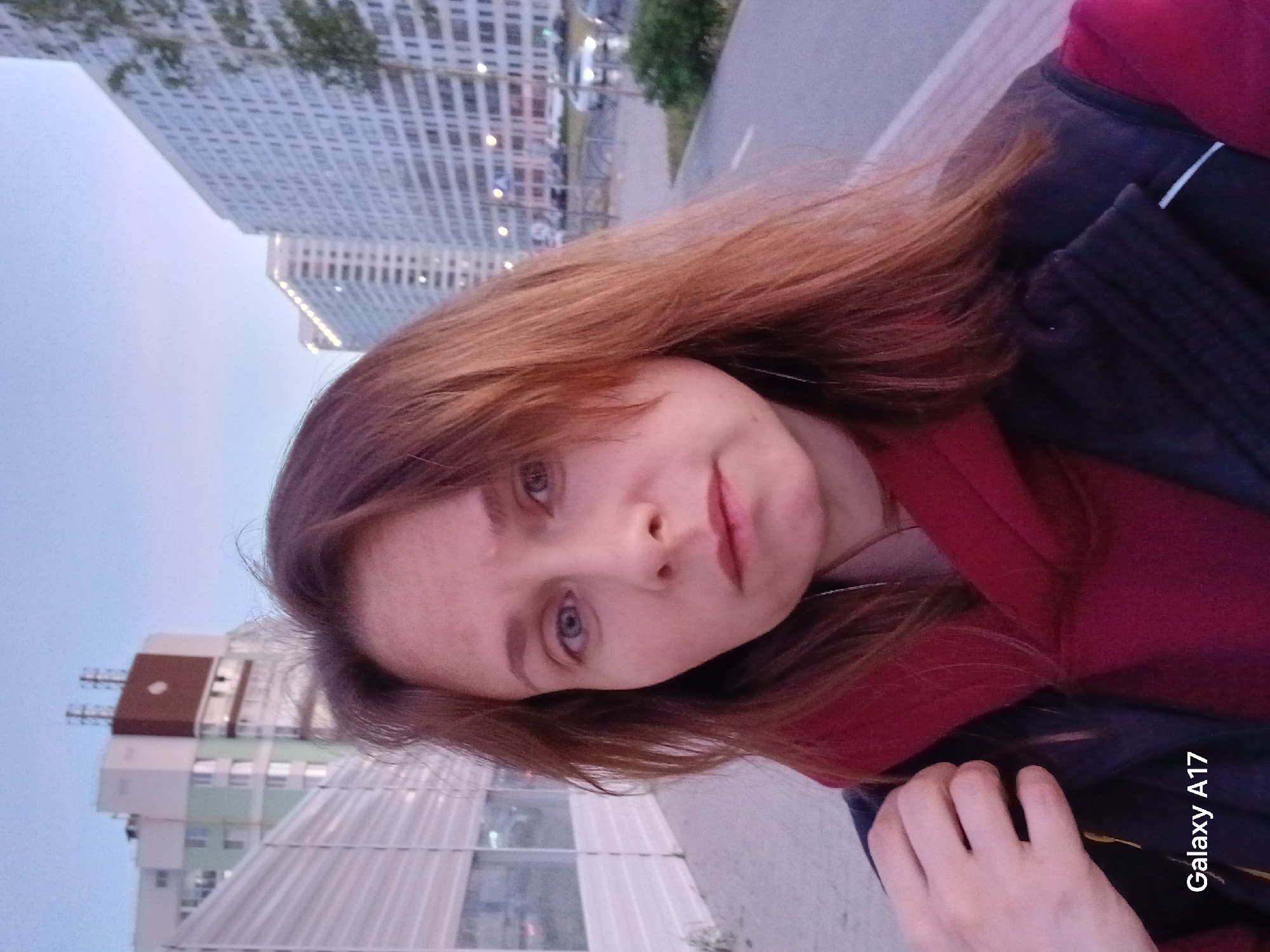 A woman is posing for a selfie in Yekaterinburg, Russia. The background features urban architecture, including tall apartment buildings. The woman has light skin, light-colored eyes, and reddish-brown hair. She is wearing a dark-colored jacket over a red top. Her hand is visible near the bottom right corner of the image. The text "Galaxy A17" is displayed on the lower right side of the image, indicating the device used to capture the photo. The overall scene suggests an urban setting during the early evening or late afternoon.