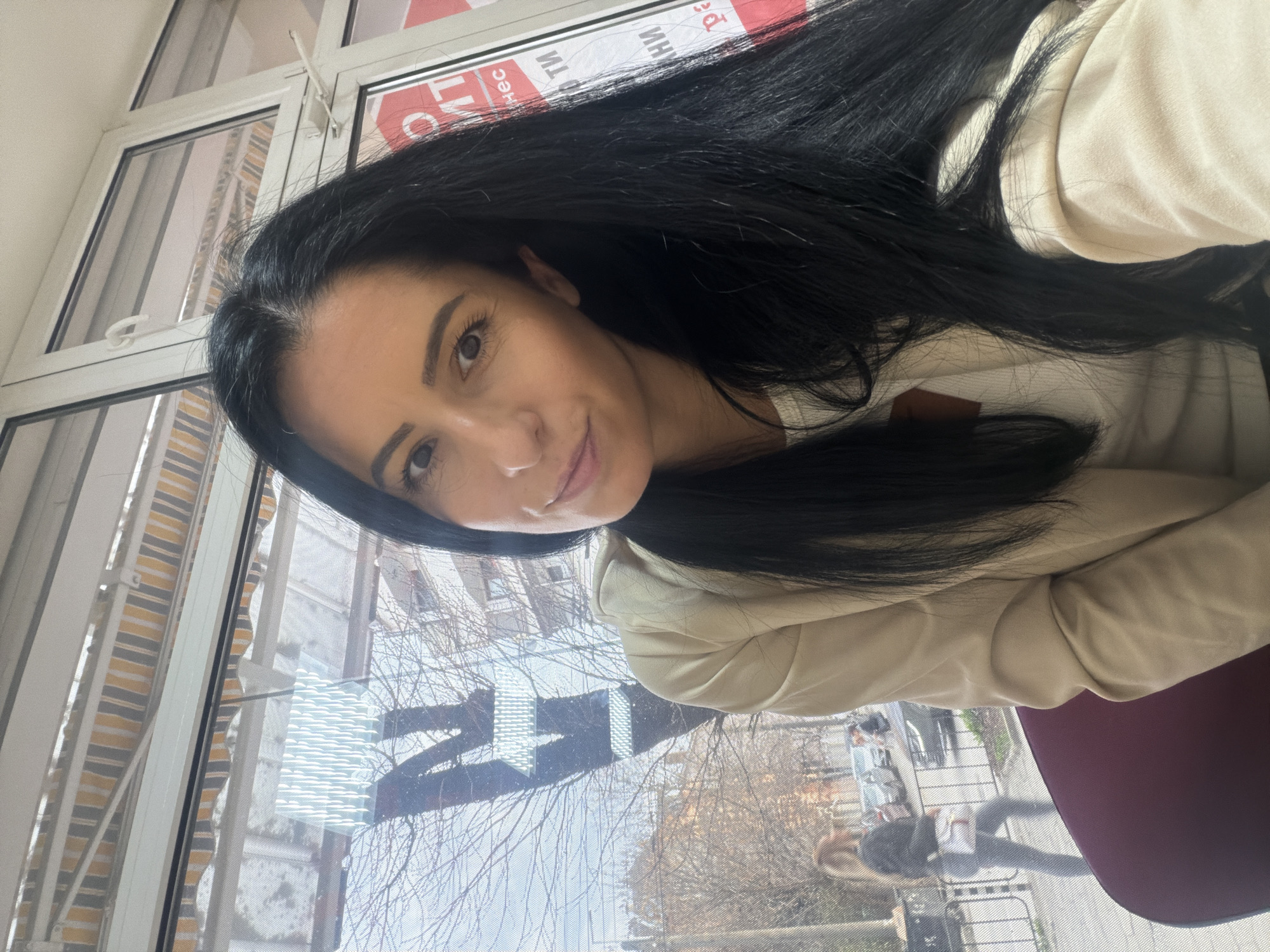 A dark-haired woman is positioned in the foreground, facing the camera with a slight smile. She has long, straight, dark hair and is wearing a light beige hooded jacket over a white garment. She appears to be seated, with a dark red chair back visible below her right arm.
She is indoors, in front of a multi-pane white-framed window with a visible screen mesh. Through the upper left portion of the window, a yellow and grey striped awning is visible. On the right side of the window, partially obscured by the woman's hair and window frame, red and white signage with Cyrillic text is visible, including fragments such as "ТИМ," "нес," "НИ," and "ра," consistent with Bulgarian language.
Outside the window, a city street scene is visible under a bright sky. Bare-branched trees line the street, suggesting a dormant season. Buildings are visible beyond the trees. A pedestrian wearing a dark top, light pants, and carrying a white bag is walking left-to-right on a sidewalk behind a black metal railing. Several parked cars are visible on the street. The scene is located in the city of Pleven, Bulgaria.