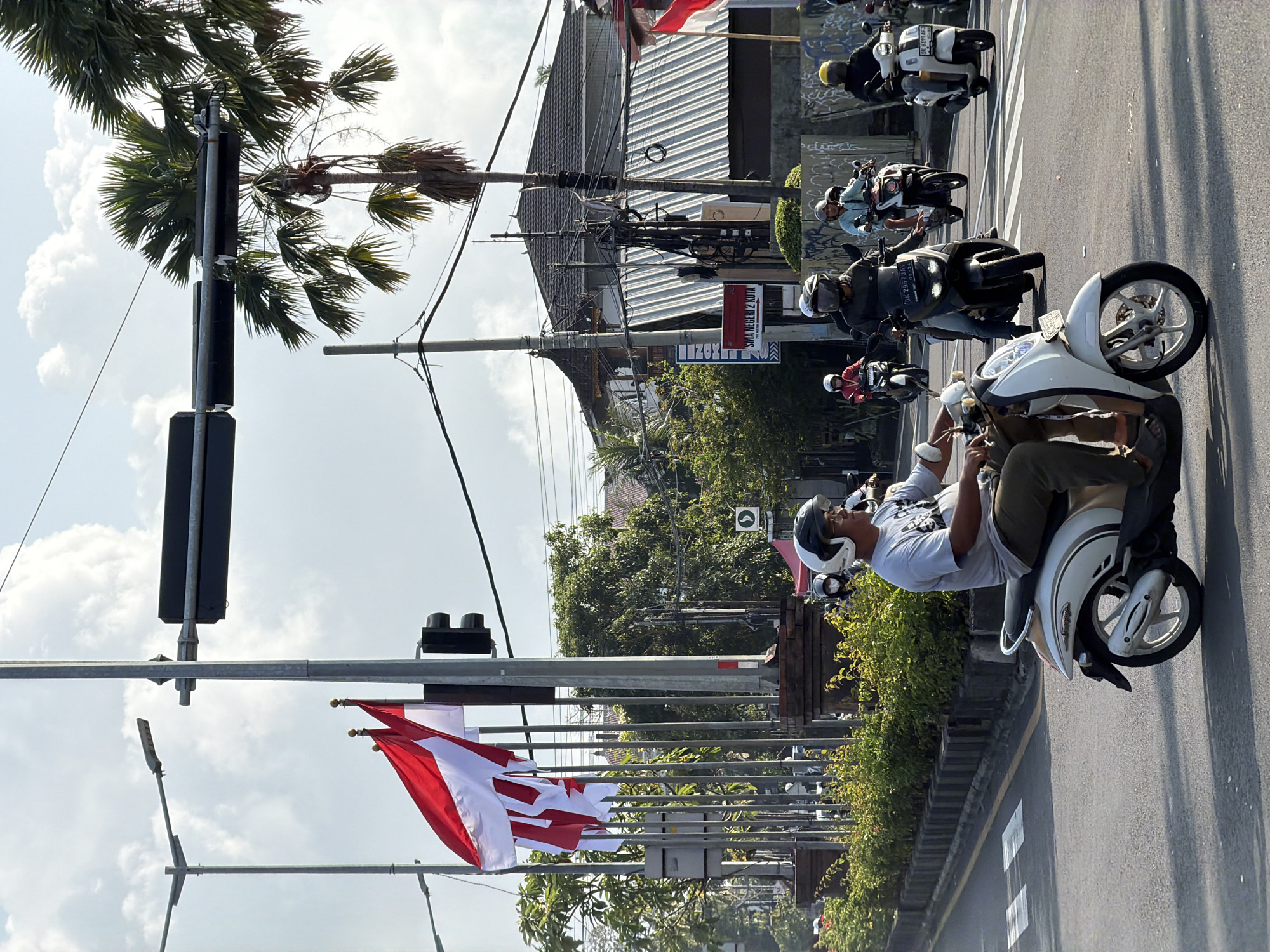 The image captures a street scene with multiple motorcycles navigating a roadway. In the foreground, a person on a white scooter, wearing a white helmet and a light-colored t-shirt, is riding on the right side of the road. Several other motorcyclists follow, some in helmets and others without. Indonesian flags, red and white, are prominently displayed on flagpoles along the roadside, next to a traffic light. Palm trees are visible in the background, with a building with a corrugated metal roof behind them. The sky is partly cloudy. The environment appears to be a city street with paved roads, sidewalks, and green vegetation lining the sides of the road.