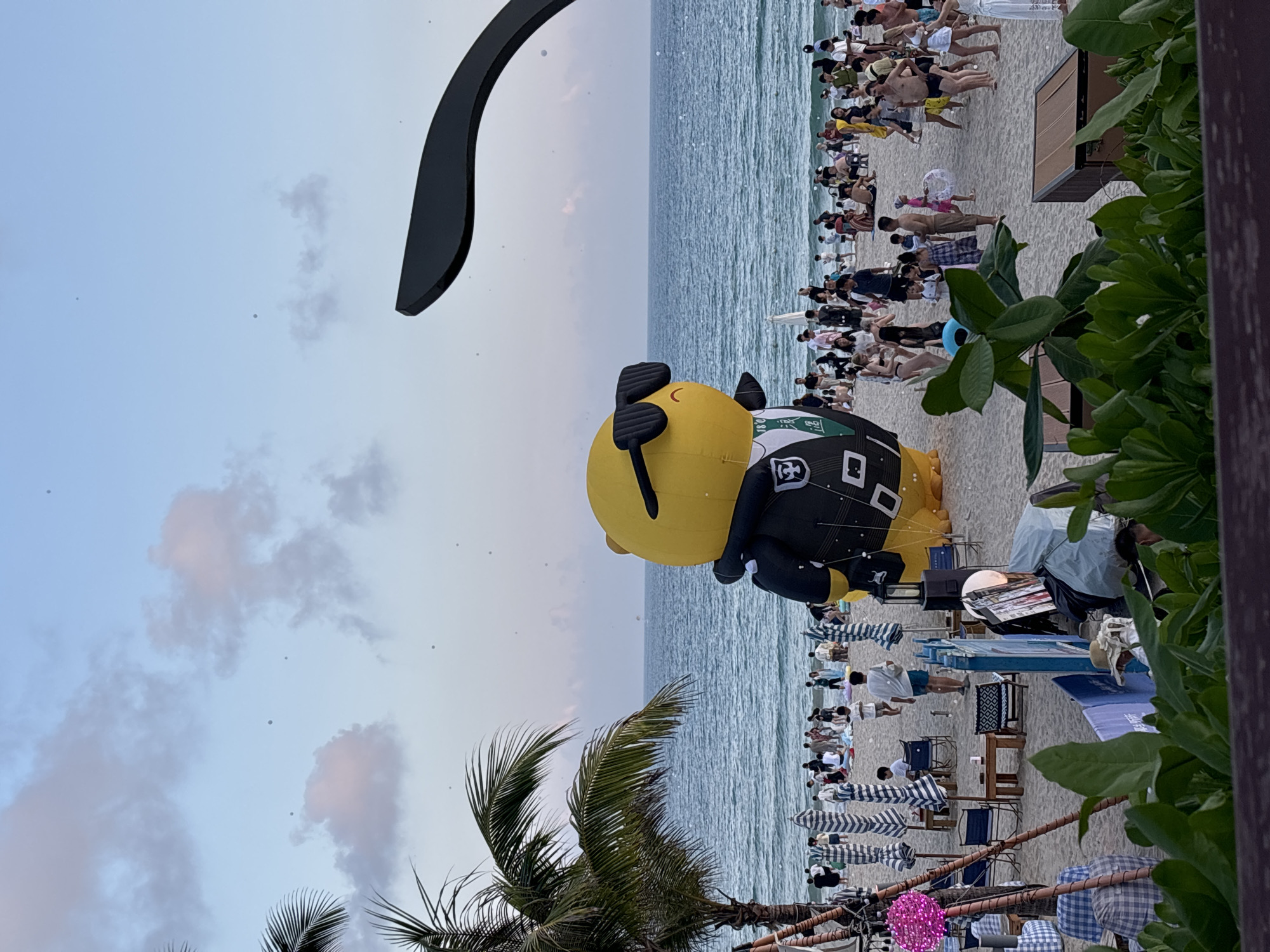 The image depicts a beach scene in the Jiyang District, China. A large, inflatable yellow character wearing sunglasses and a black suit with a green tie stands prominently on the sand. The character has a stylized, smiling face. In the background, the ocean stretches to the horizon under a pale blue sky with scattered clouds. A crowd of people are scattered across the beach, some sitting under umbrellas, others walking or standing. Beach umbrellas and furniture are visible, suggesting a recreational area. Lush green foliage is in the foreground, partially obscuring the lower part of the scene. The overall atmosphere appears to be that of a public gathering or event on a beach.