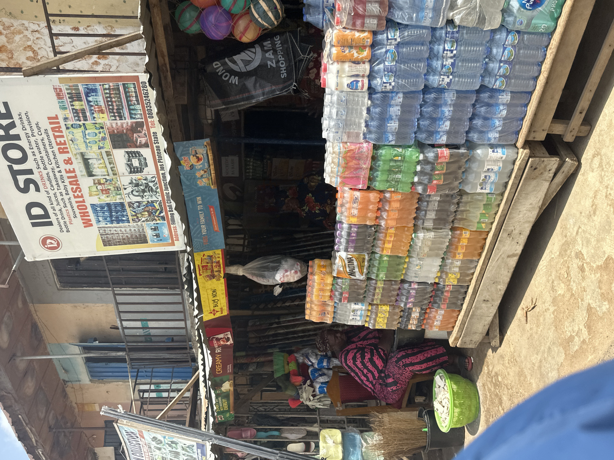This is a medium shot, captured from a slightly elevated angle, of a bustling outdoor market stall in Jos, Nigeria. The scene is characterized by an abundance of bottled drinks, arranged neatly in stacks on wooden shelves. The variety is evident, with rows of water bottles, soft drinks, and what appear to be energy drinks in various colors and packaging.
In the foreground, a woman is seated, wearing a vibrant pink and black striped garment, her legs crossed. She appears to be tending to the stall or taking a break. To her left, a green woven basket overflows with white items, possibly packaging or small goods. Behind her, a display of brooms and other cleaning supplies is visible, adding to the diverse inventory of the stall.
Above the bottled drinks, several signs and banners are displayed. One prominent sign, oriented vertically, reads "ID STORE" and details the types of products sold: "Energy Drinks, Bottle Drink, Juices, Candies, Bottle water, Provision, Plastics Such as Takeaway, Containers, Cups, Souvenirs, Baby Kits & Kitchen Utensils." Other signs include "BUY NOW!", "YOUR FAMILY TO WIN," and brand names like "MAGGI" and "pepsi." The overall impression is one of a well-stocked retail space catering to everyday needs.
The lighting suggests it is daytime, with sunlight casting shadows and highlighting the textures of the goods and the weathered wooden structures of the stall. The presence of multiple brands and the variety of items point to a busy commercial environment. The scene evokes a sense of everyday commerce and community interaction typical of a Nigerian market.