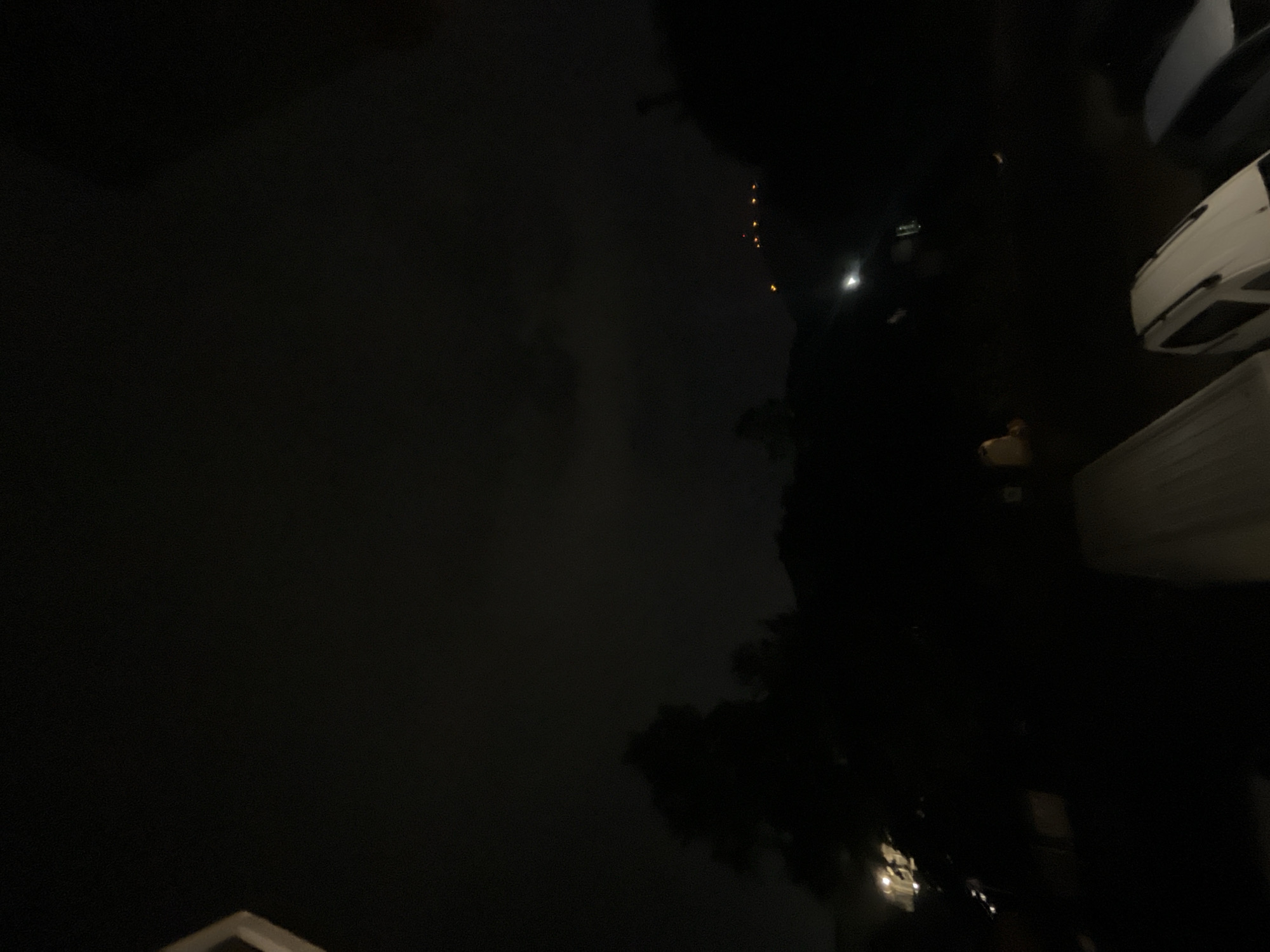 A nighttime, low-light, wide-angle shot captures a dark, silhouetted landscape with a faint, cloudy sky. In the foreground, the top of what appears to be a light-colored, parked vehicle is visible. Further in the background, several other vehicles are dimly illuminated, suggesting a parking area. Trees and a hillside are silhouetted against the sky. Distant lights, possibly from buildings or streetlamps, are visible on the crest of the hill. The scene is devoid of people. The overall impression is one of a quiet, possibly suburban or semi-rural area at night. The specific location within Plovdiv, Bulgaria, cannot be determined from the visual information provided.