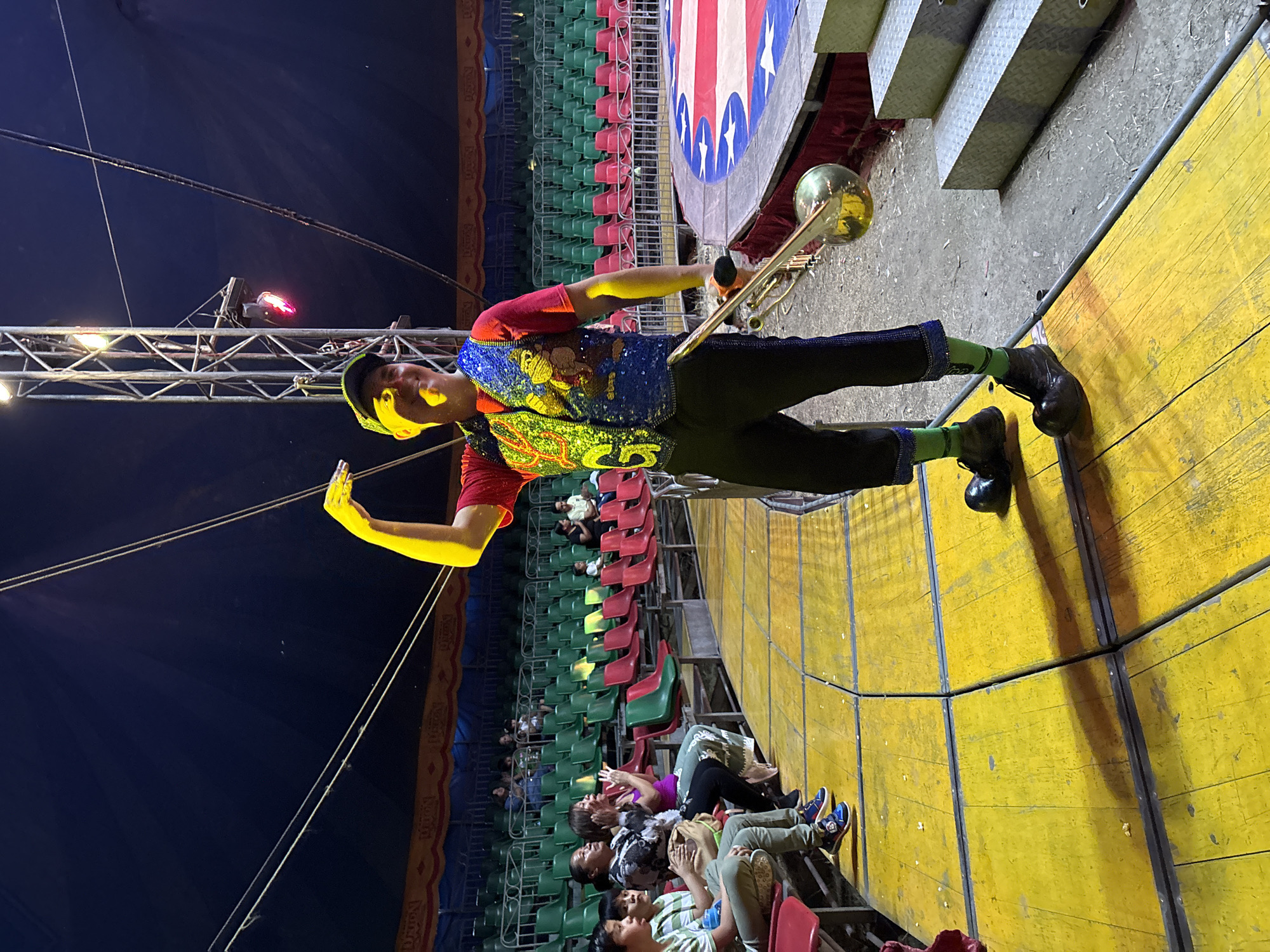 Inside a circus tent in Tejalpa, Mexico, a performer stands on a yellow platform, dressed in a red shirt, sequined vest, dark pants, and green socks, holding a trombone. Audiences fill red and green seats facing the stage. A light structure and part of the blue circus tent roof are visible. A platform with a stars-and-stripes backdrop sits nearby.