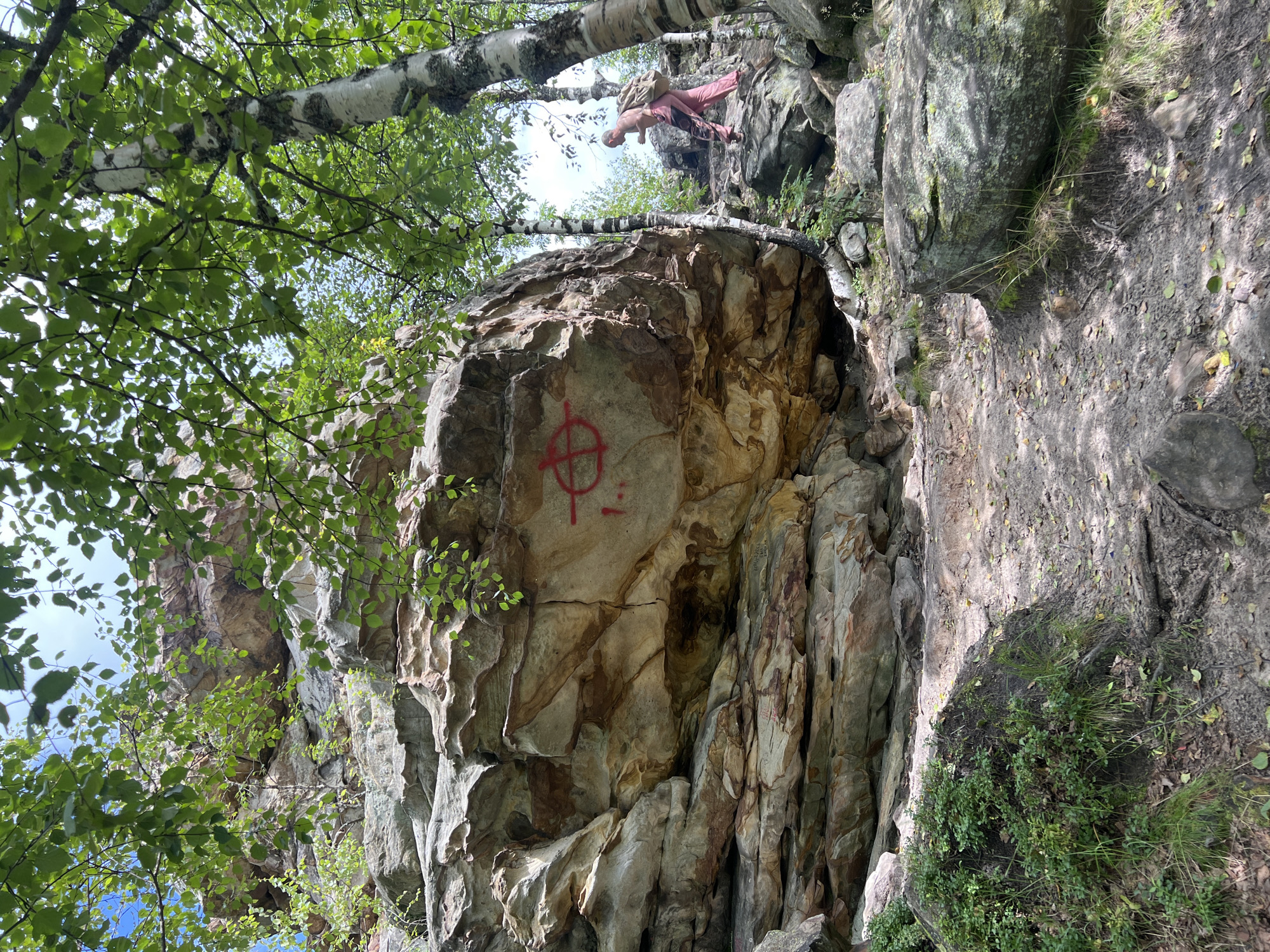 A person wearing pink pants and a backpack is walking up a rocky trail near a large rock formation. A red symbol, resembling a circle with a cross and two horizontal lines, is painted on the rock face. The scene appears to be in a forested area with birch trees. The location is near the city of Gubakha, Russia.