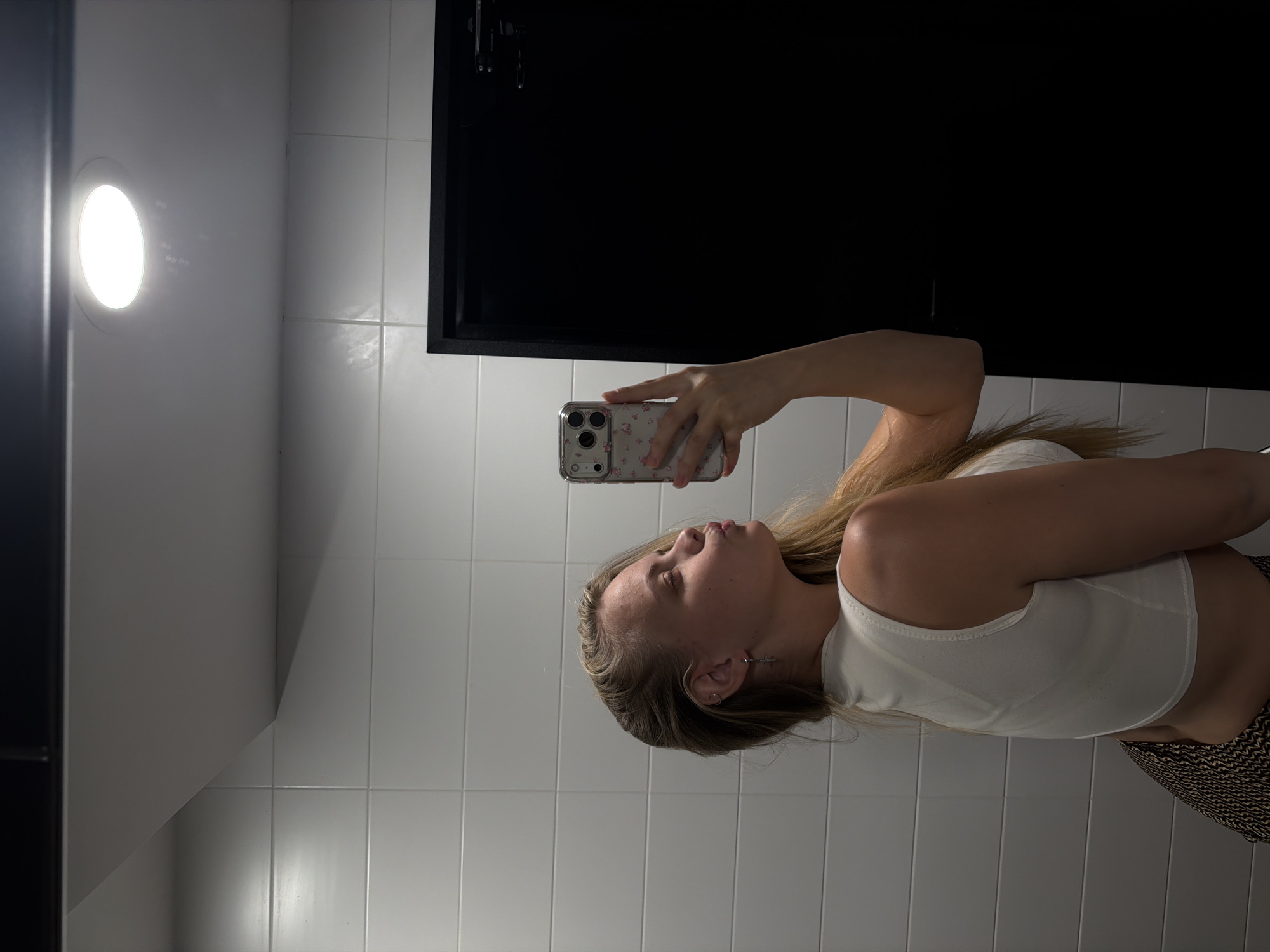 A person is taking a selfie in a bathroom with white tiled walls. A bright round light fixture is visible in the upper left corner, and a black framed structure, possibly a mirror or cabinet, is positioned in the upper right. The person is holding a smartphone with a floral case and wearing a white tank top. The background consists of white tiles. There is no evidence suggesting this is Izhevsk, Russia.
