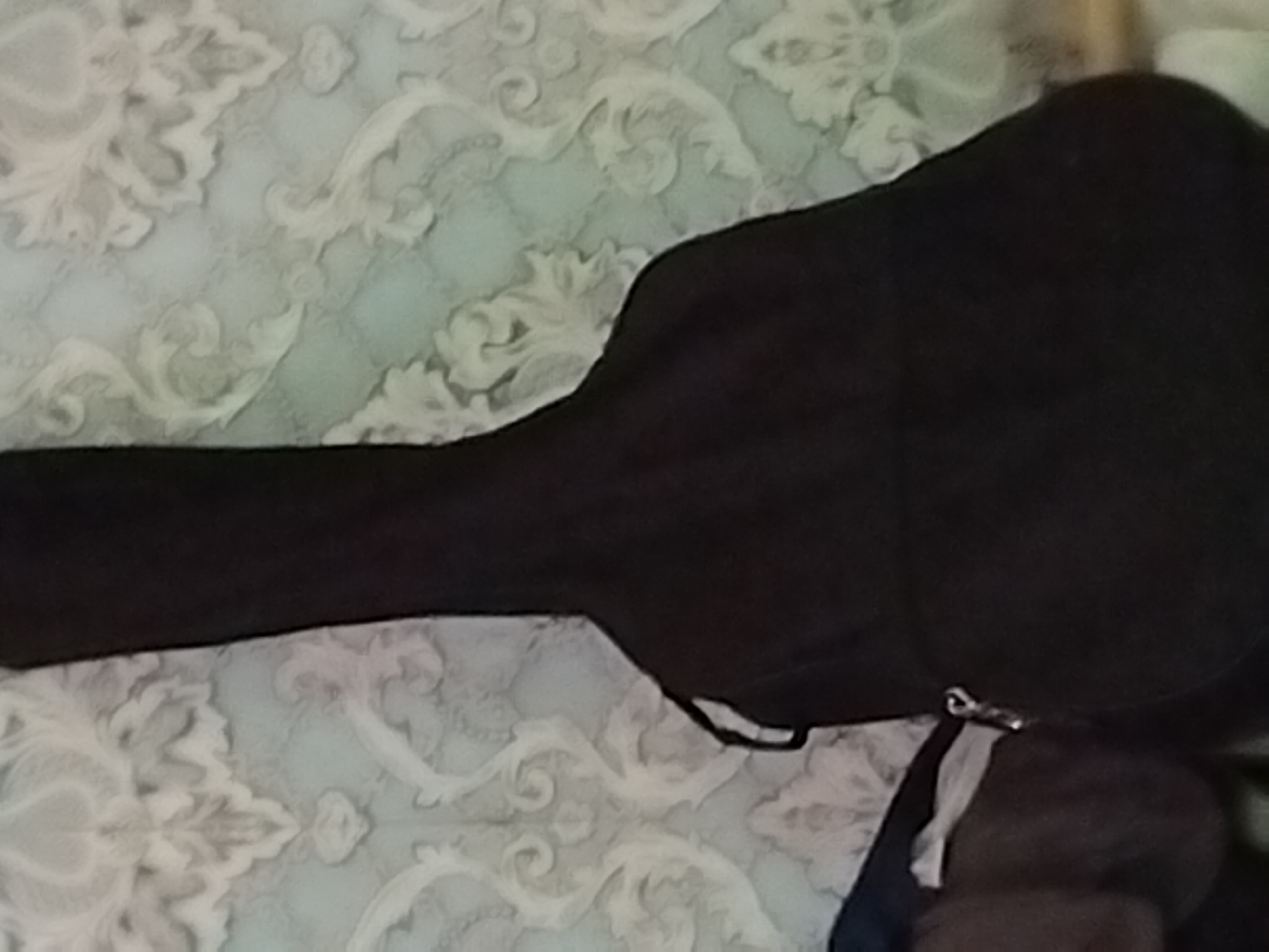 A black guitar case is leaning against a wall with ornate, light-colored wallpaper. The case is oriented vertically and appears to be made of fabric. A small metal zipper pull is visible on the side of the case. The environment suggests an indoor setting, possibly a room in a residential or storage area within Pervozvanivka, Ukraine. The overall lighting is somewhat dim.