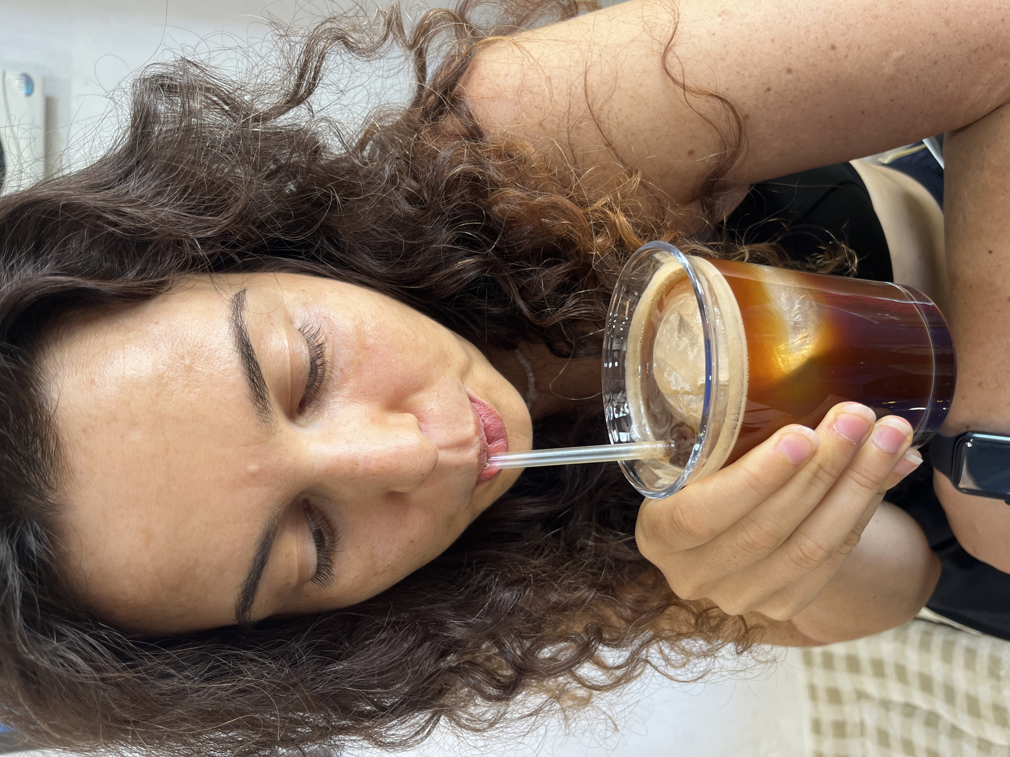 The image shows a close-up of a woman with curly brown hair drinking an iced coffee from a clear plastic cup. She is using a white straw and her eyes are closed. The coffee is dark brown with a layer of foam and several ice cubes. The woman is wearing a black tank top. The background is blurred but appears to be indoors with a light-colored wall. There is no information in the image to indicate a specific location in Phu Thuy Ward, Vietnam.