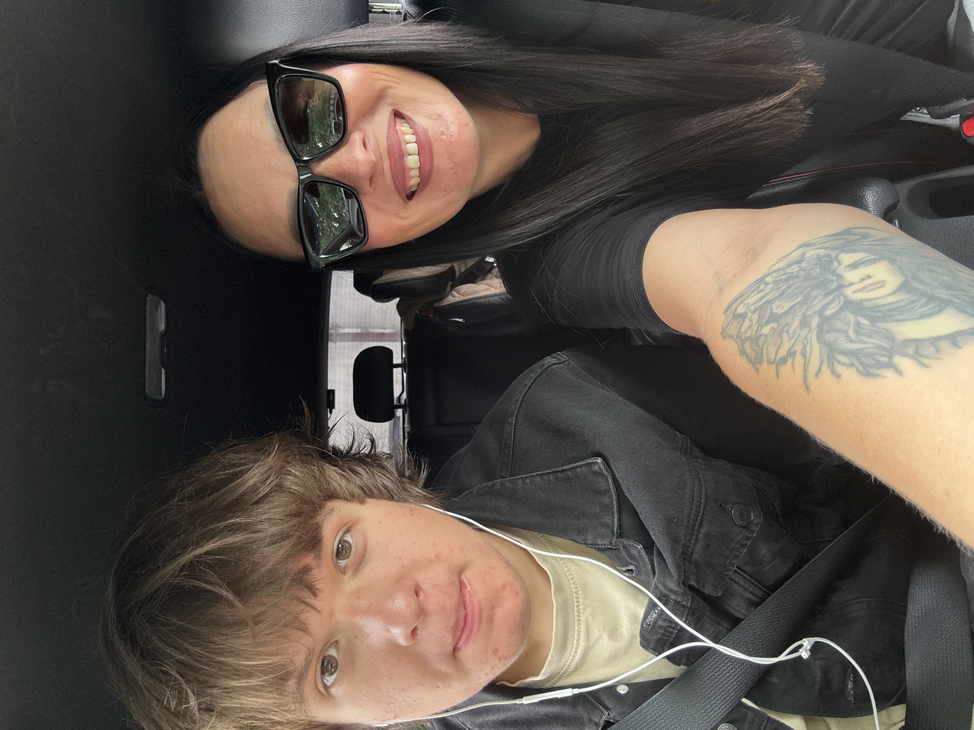 This is a selfie taken inside a vehicle, likely a car, given the presence of a seatbelt and car interior elements. The image is oriented sideways.
The foreground features a young person with light brown, somewhat messy hair and light brown eyes. They are wearing a beige t-shirt and a dark denim jacket. Earbuds are visible, with the white cord running across their chest. The person appears to be looking directly at the camera with a slight smile. Their skin has some blemishes and freckles.
In the background, a woman with long, dark hair and dark-rimmed sunglasses is also visible. She is smiling broadly, showing her teeth. She is wearing a black top. A tattoo of a woman's face is visible on her arm. The interior of the car is dark, with hints of a black dashboard and console. The overall impression is of a casual moment captured between two people. The lighting suggests it's daytime, possibly late afternoon or early evening, given the slightly dimmer interior lighting compared to the outdoors reflected in the sunglasses.