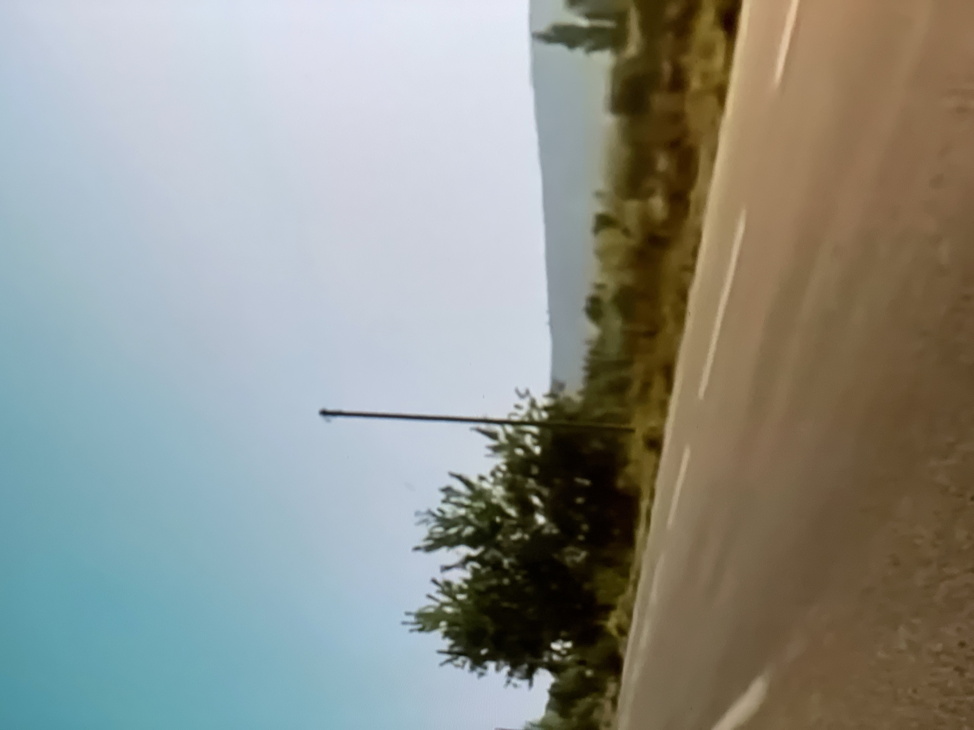 The image depicts a highway curving to the right, bordered by sparse vegetation and hills in the background. The sky is a pale blue, suggesting daytime. A utility pole stands on the left side of the road, near some greenery. The scene appears to be a rural or semi-arid environment, possibly in a location like Aktau, Kazakhstan, given the context. There is no text visible in the image. The overall impression is one of a journey or a drive through a somewhat desolate landscape under clear skies.