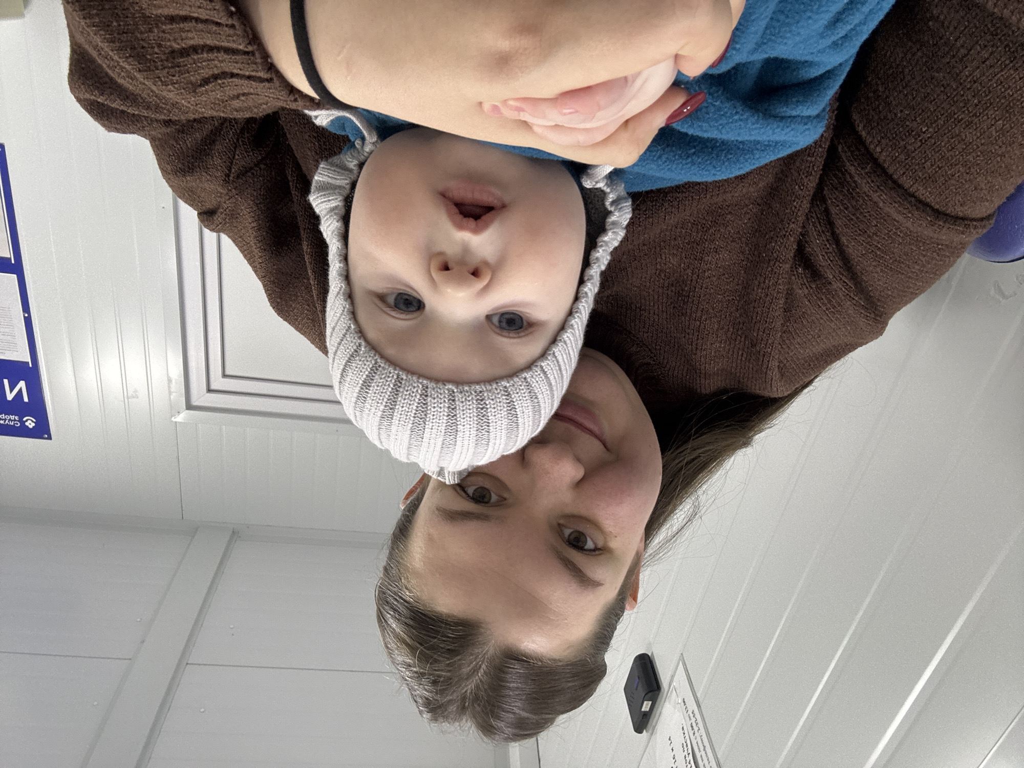 A woman and a baby are positioned closely together, facing the camera. The woman has brown hair and is wearing a brown sweater. The baby is wearing a light gray knit hat and a blue top. A hand with red nail polish is visible holding the baby. The background consists of white wall panels. A sign in Cyrillic script is visible on the wall. The location is Марковский, Russia.