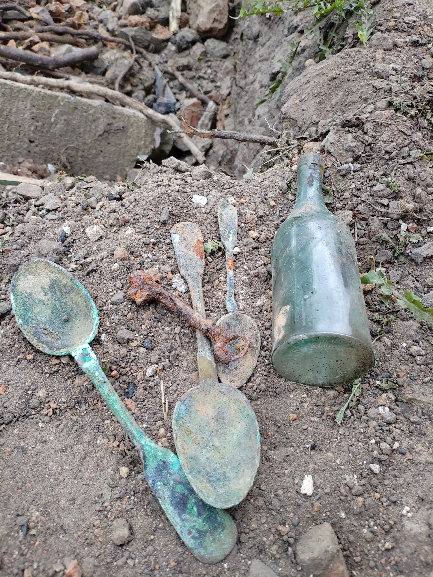 An outdoor, ground-level, medium shot captures an assortment of aged artifacts partially embedded in dark soil and small stones. In the foreground, arranged from left to right, are four tarnished spoons. The two largest spoons on the left exhibit a patina of green and black, indicative of significant corrosion or age, with one spoon’s bowl appearing to be made of a different material or heavily damaged. To the right of these, two smaller, distinctly rusted spoons are clustered together, one with a corroded handle twisted and exhibiting signs of metal decay. Adjacent to the spoons, and slightly further back, is a tall, dark green glass bottle, upright and showing signs of wear and dirt accumulation. The background consists of a mix of soil, larger stones, and debris, including what appears to be a piece of concrete or brick. The overall environment suggests a site of excavation or accumulated refuse, possibly in a rural or unmaintained area. The specific location is identified as Bialynichy, Belarus.