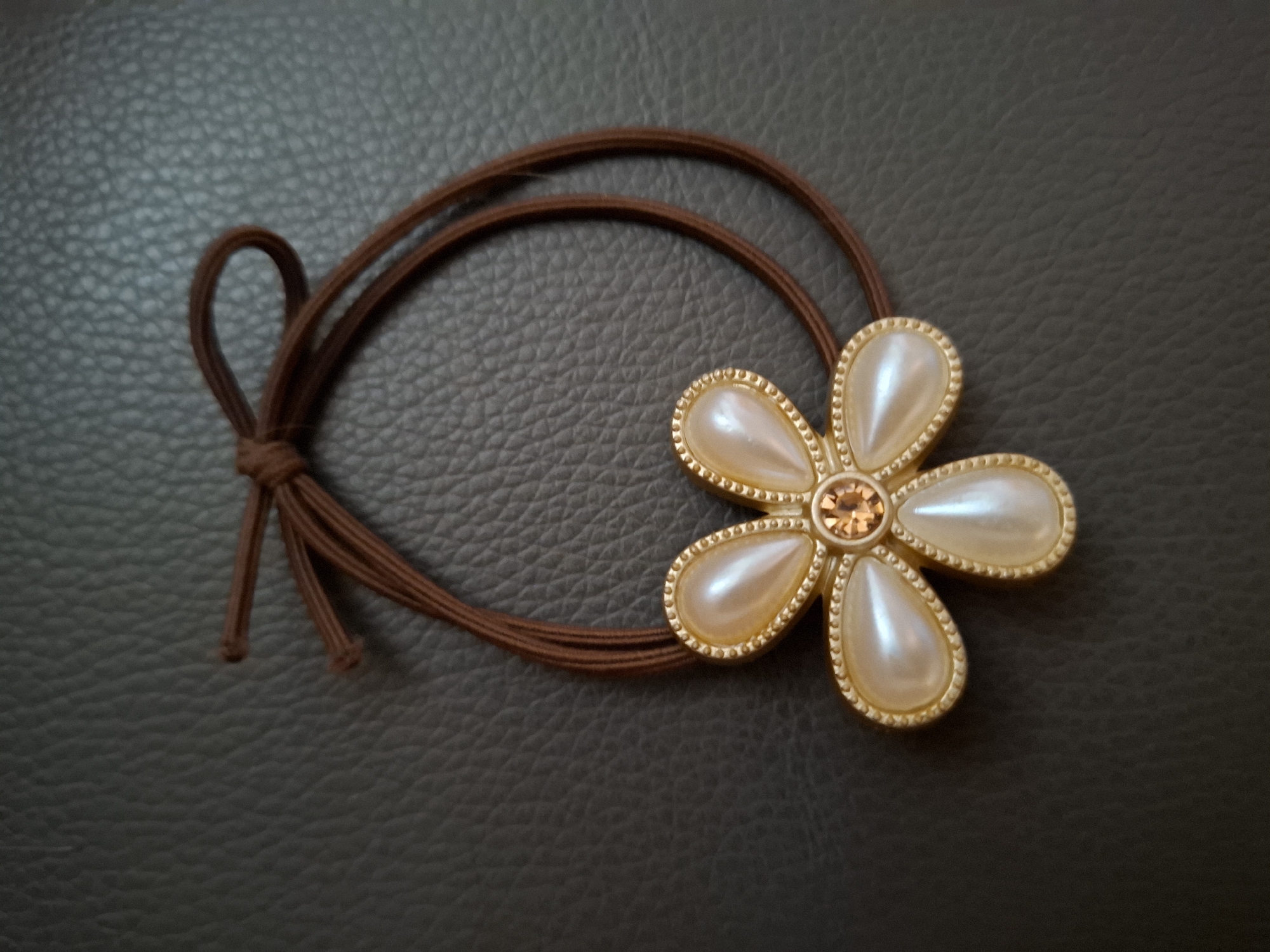 A close-up, eye-level, horizontal shot shows a brown hair tie with a decorative flower-shaped embellishment lying on a dark, textured surface. The hair tie itself is a simple, thin, dark brown elastic band that has been tied into a small knot, with two short ends dangling from it. The flower embellishment is attached to the elastic band and faces to the right. It has five teardrop-shaped "petals" that appear to be made of a pearlized material in a creamy white color. Each petal is outlined with a thin, textured gold trim that has small raised bumps along its edge. In the center of the flower is a small, round, faceted gemstone in a light amber or peach color. The dark surface beneath the hair tie has a pebbled or grained texture, resembling leather or a similar material. The lighting suggests an indoor setting, possibly with artificial light, as there are no distinct shadows indicating natural sunlight. The image is a still life, focusing solely on the object and its immediate surroundings. No people, animals, or text are visible in the frame. The overall mood is neutral and focuses on the aesthetic details of the accessory.