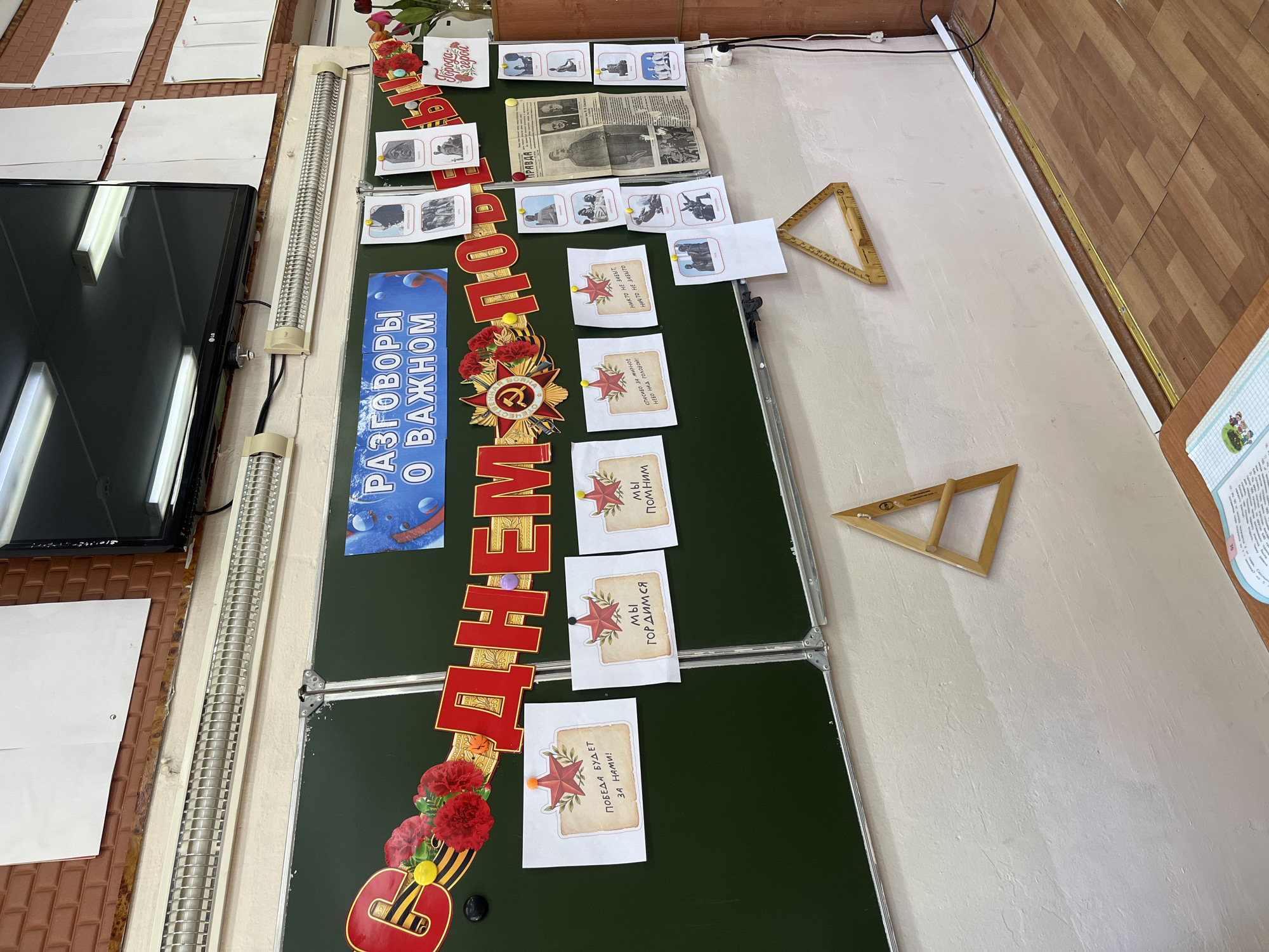 The image captures an indoor setting, likely a classroom or meeting room, featuring a green chalkboard adorned with decorations and educational materials related to Victory Day in Russia. The chalkboard is decorated with red lettering spelling out "Victory Day," accompanied by imagery of red flowers, Soviet medals, and stars. The phrase "Conversations About Important Things" is printed vertically on a blue banner. The board also displays pictures of war events, newspaper clippings, and printed cards with a red star and slogans such as "We remember", "We are proud", and "Victory will be ours." There are two wooden triangles, possibly for mathematical use, affixed to the right side of the board. The walls are painted white, with a brown wooden panel to the right. A flat-screen TV is mounted on the wall to the left, alongside fluorescent lighting fixtures. Other details include blank sheets of paper pinned to the wall near the TV and a blurred view of a desk and a printed poster on the right side. The photograph appears to have been taken in Kaspiysk, Russia, based on the text displayed and the Victory Day theme, which is of national significance in Russia.
