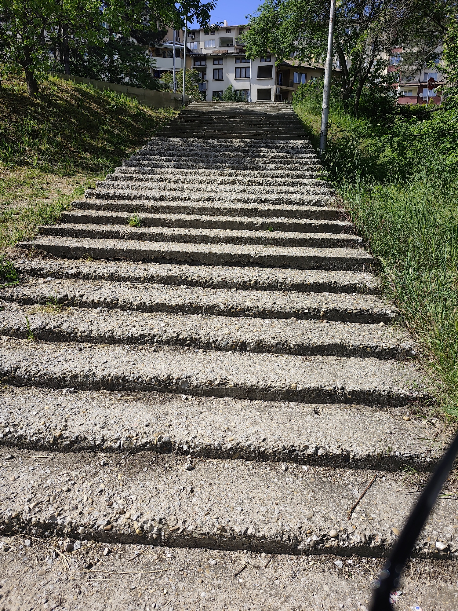 The image depicts a long, concrete staircase leading uphill towards a building. The steps are worn and slightly overgrown with grass and weeds on the sides. To the left of the staircase is a grassy slope with trees, while to the right there is taller grass and bushes. The building at the top of the stairs appears to be a residential apartment complex. The location is Unknown, Unknown. A black shadow is cast onto the stairs in the foreground.