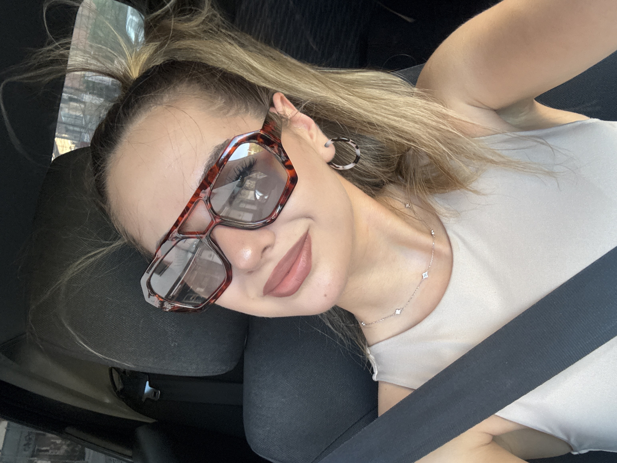 A woman is in a car, wearing glasses, a light-colored top, and a seatbelt. She has blonde hair pulled back into a ponytail. She is wearing hoop earrings and a necklace. The location appears to be inside a vehicle, possibly in Blagoevgrad, Bulgaria. However, specific location information is not discernible from the image.