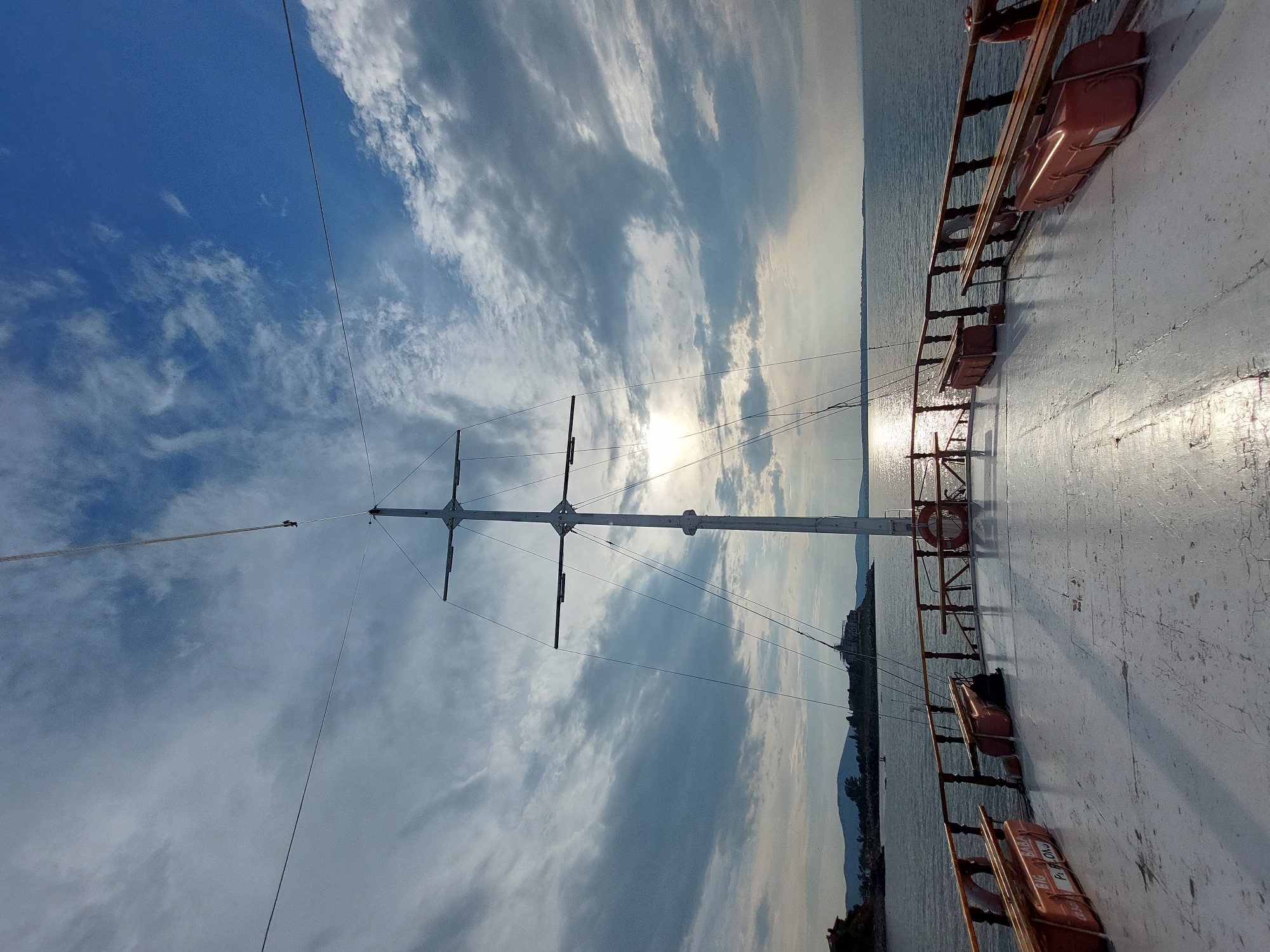 The image depicts the deck of a boat, likely a wooden sailing vessel, viewed from a low angle looking forward. A prominent mast with several spars dominates the center of the frame, silhouetted against a bright, partially cloudy sky. The sun is visible, glinting off the water. The deck is weathered and painted white, with wooden railings along the sides. Orange life preservers are attached to the railings. In the distance, a coastline with hills and some buildings is visible. The sea is calm. This scene is consistent with the waterfront area of Sozopol, Bulgaria, a historic coastal town known for its maritime heritage and picturesque harbor.