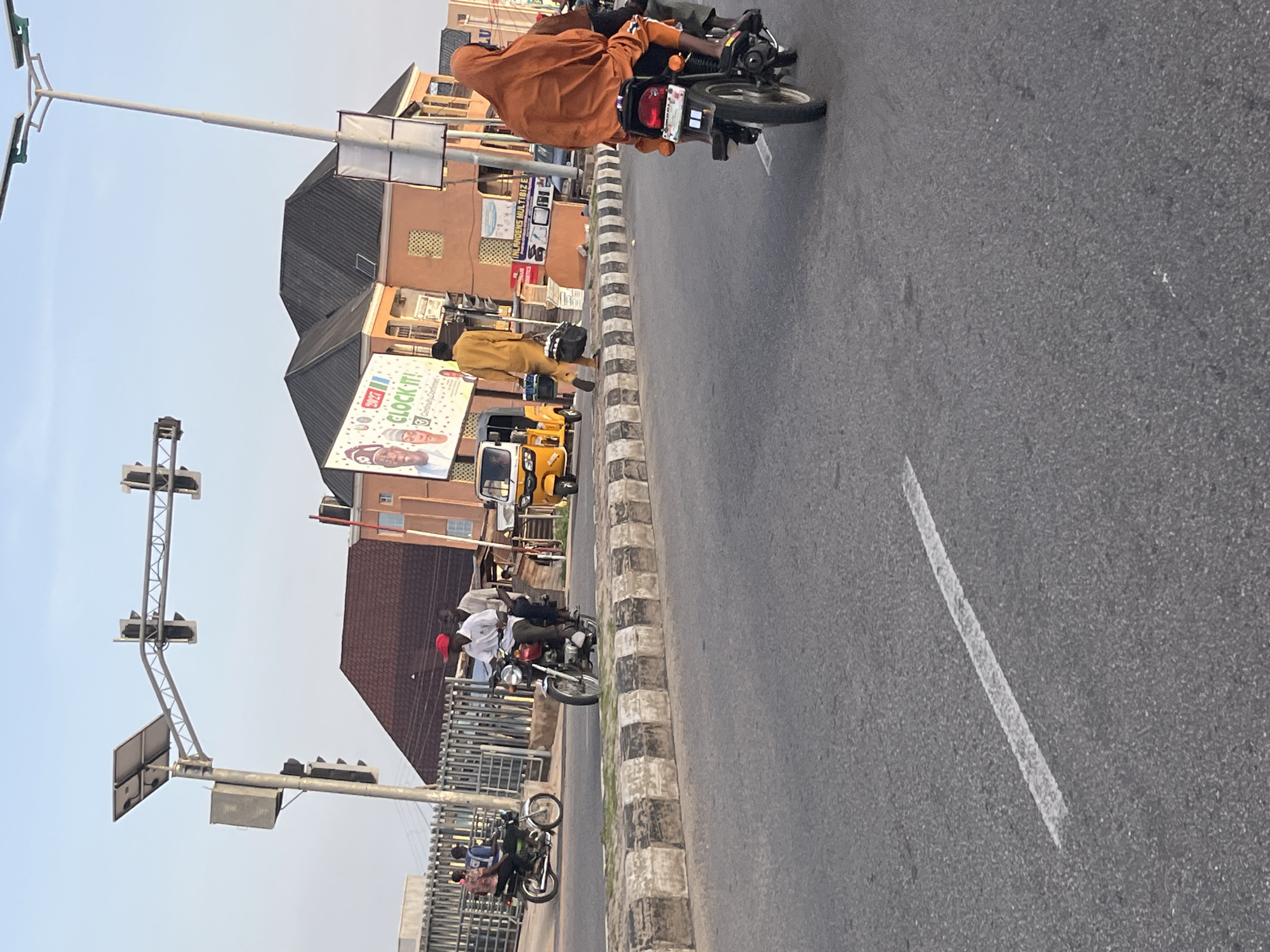 The image captures a street scene in a city with a bright, clear sky. Several motorcycles are in motion. On the right, a person wearing an orange headscarf and garment rides a motorcycle. In the middle ground, two people are riding a motorcycle, with the rider in front wearing a red cap and white shirt. To the left, another motorcycle carries two individuals. In the background, a yellow auto-rickshaw is parked or moving slowly. Buildings with orange and brown facades are visible, one featuring a large advertisement with the text "CLOCK-IT!" and portraits. Traffic lights and solar panels are mounted on poles overhead. The road is paved, with a white line marking a lane. A concrete barrier with black and white stripes separates the road from a sidewalk or pedestrian area. The overall impression is a bustling urban environment with active transportation.