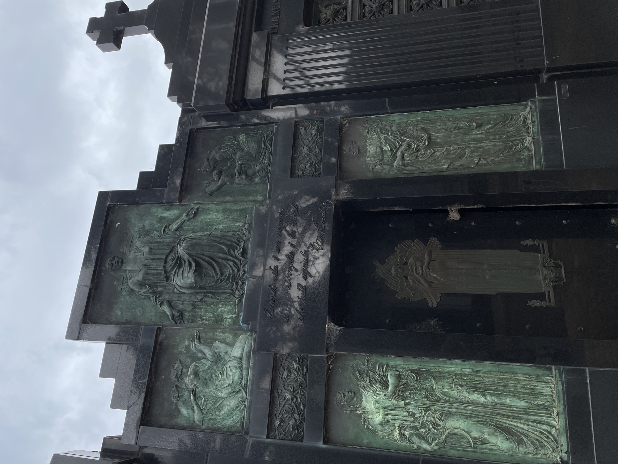 A large, dark, ornate mausoleum stands under an overcast sky in the Recoleta Cemetery, Buenos Aires, Argentina. The structure is primarily constructed from dark, polished stone, possibly granite, with a stepped roofline on the left and a prominent, unadorned cross on the right. The facade is extensively decorated with multiple bronze reliefs, all exhibiting a distinct verdigris patina.
A large central upper relief depicts a seated, draped female figure with outstretched arms. To its left, a relief shows two or three draped figures, one kneeling. To the right, another relief features a figure tending to or embracing a smaller figure. Below these, horizontal decorative panels display intricate floral or foliage motifs.
The central entrance is a dark metal door, featuring a tall, vertical bronze relief of a winged figure holding an object to its chest. A small, oxidized metal lock is attached to the right side of this door. Above the door, an inscription in Italian is visible, reading: "Questa vita è un sogno, il risveglio è la morte che tutti aspettiamo. Vivere è morire." (This life is a dream, the awakening is death that we all await. To live is to die.)
To the lower left of the door, a tall bronze panel portrays a draped female figure holding a sheaf of wheat. To the lower right, another panel shows two standing, draped figures. The far right section of the mausoleum incorporates fluted pilasters and intricate metalwork grilles. Sparse cobwebs are visible on the stone surfaces, particularly around the inscription. No individuals are present in the scene.