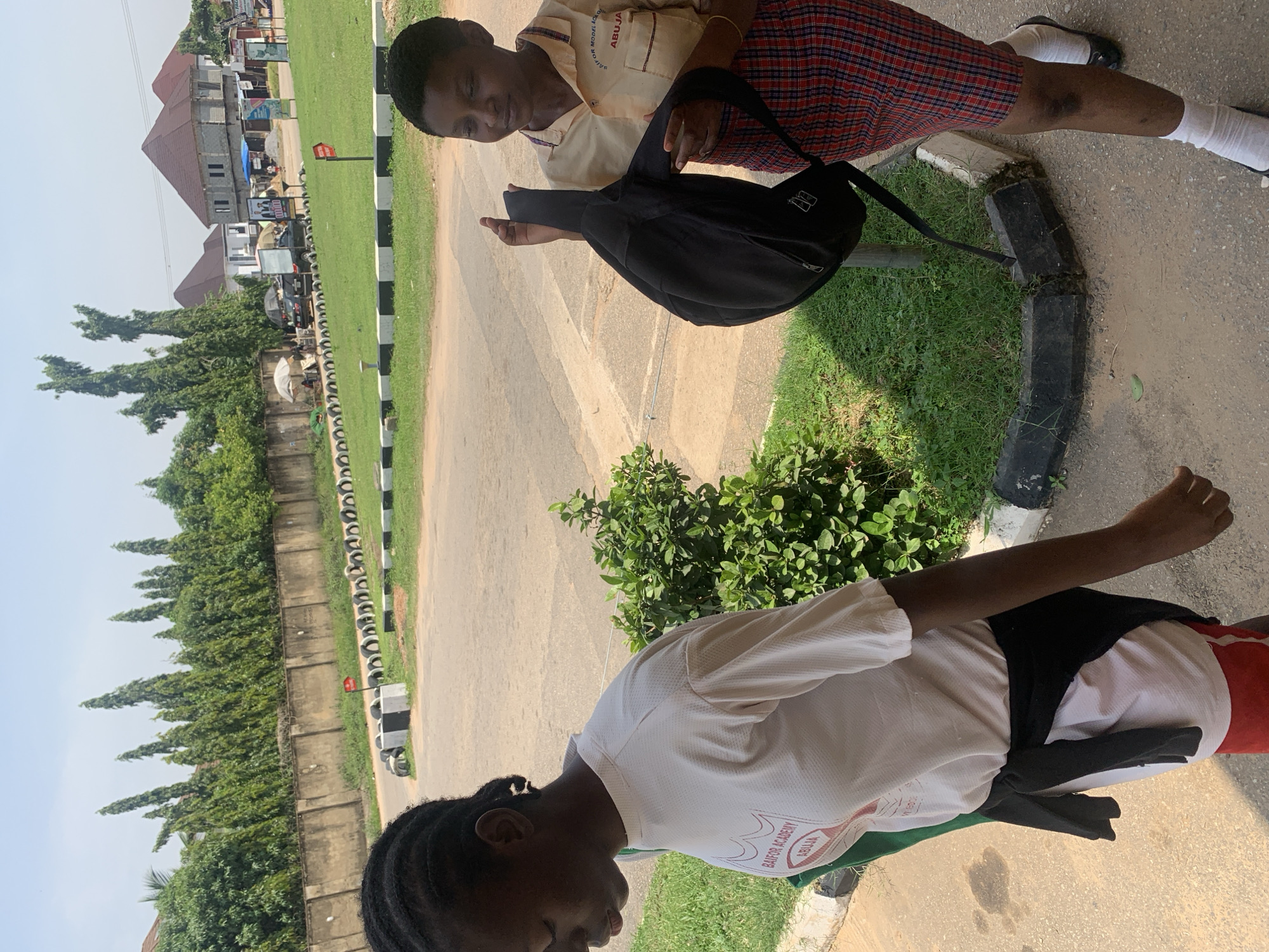 Two young Black females are walking outdoors on a paved surface near a grassy area with a line of black and white tires serving as a border. The female on the left, with dark braided hair, wears a white t-shirt with a logo that reads "BAIFOR ACADEMY ABUJA" in red and white text. She also has a black sweater tied around her waist and is carrying a green item over her shoulder. The female on the right, with short dark hair, wears a light yellow short-sleeved collared shirt with a similar logo and "ABUJA" written on it, along with a red and blue plaid skirt. She is holding a black backpack with both hands, appearing to adjust it. In the background, there is a concrete wall, a row of tall, slender trees, and some buildings with red tiled roofs. The sky is bright and overcast. The scene appears to be a school environment in Abuja, Nigeria, given the visible signage.
