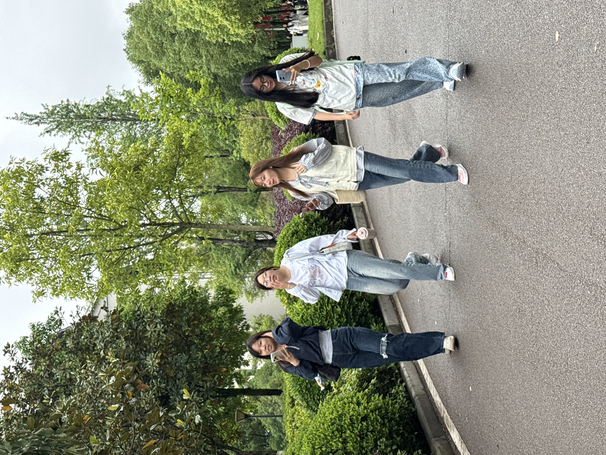 Four young women are walking together on a paved pathway in a park-like setting. The setting includes trees and manicured shrubbery. The sky is overcast. The young women are spaced apart and appear to be engaged in individual activities with their mobile phones. The location is Jiangjin, China.