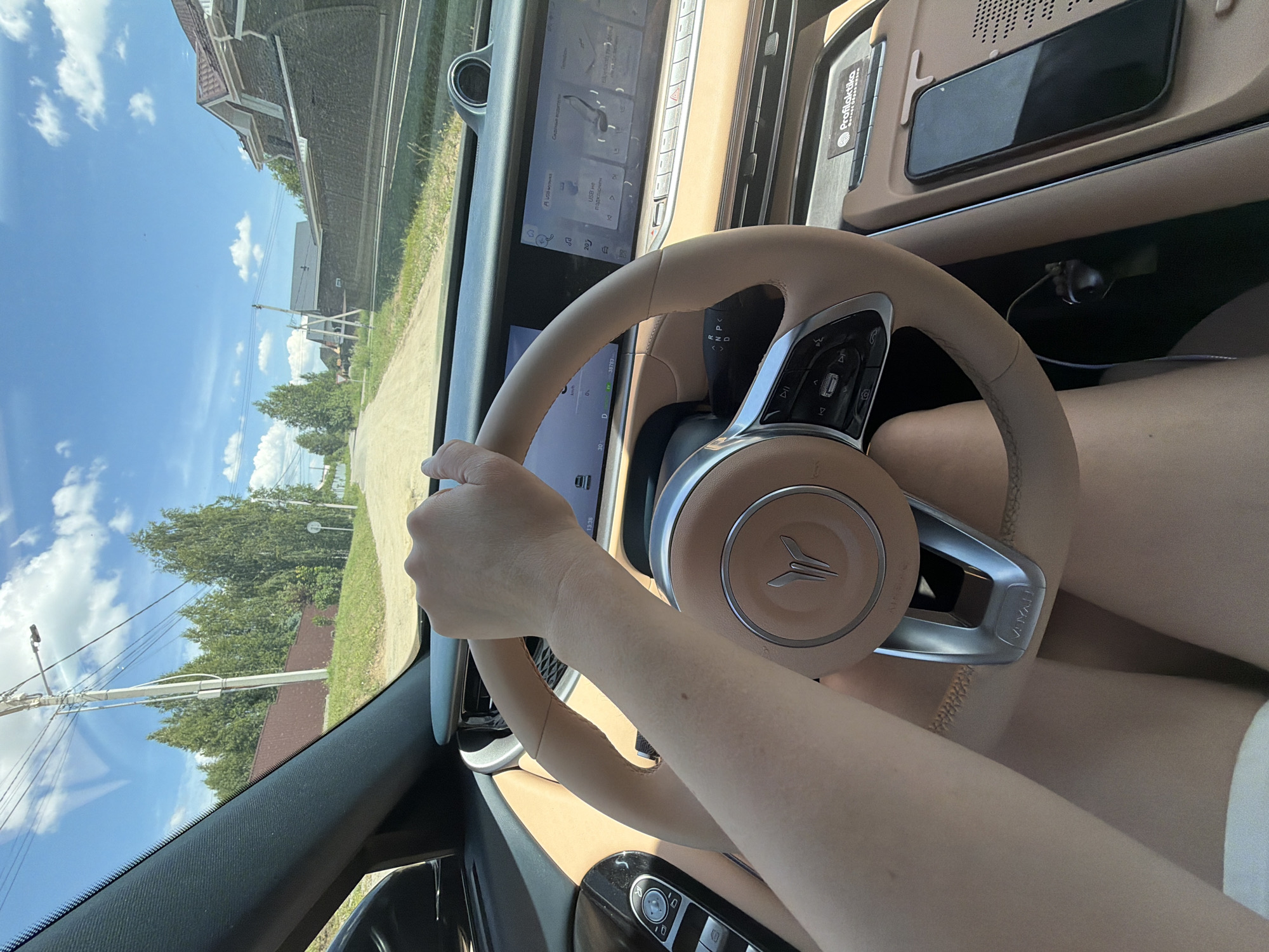 A first-person perspective captures a person driving a car on a sunny day, with the exterior view visible through the windshield. The scene appears to be a rural road, likely in Malinovka, Russia, judging by the presence of wooden fences, modest houses with pitched roofs, and lush green trees lining the roadside. The road itself is unpaved and dusty. Inside the car, the steering wheel, dashboard, and a large touchscreen display are prominent. The driver's right arm is visible, resting on the steering wheel. A smartphone is placed on the center console, indicating it's within the car's cabin. The overall impression is one of a casual drive through a suburban or rural setting.