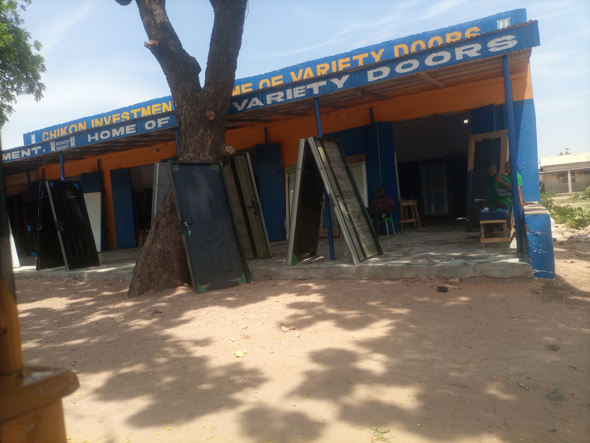 The image depicts the exterior of a business establishment in Arawa, Nigeria, identified by signage as "CHIKON INVESTMENT HOME OF VARIETY DOORS." The building features a blue facade with an orange awning. Displayed outside are numerous doors of various styles, leaning against the building and on the ground. Several individuals are visible, including a man seated inside the establishment, and two people, a woman and a man, standing near the entrance. The ground is covered in dirt and sand, with dappled sunlight filtering through tree branches, suggesting an outdoor, possibly open-air, setting. The overall scene conveys a commercial environment dedicated to the sale of doors.