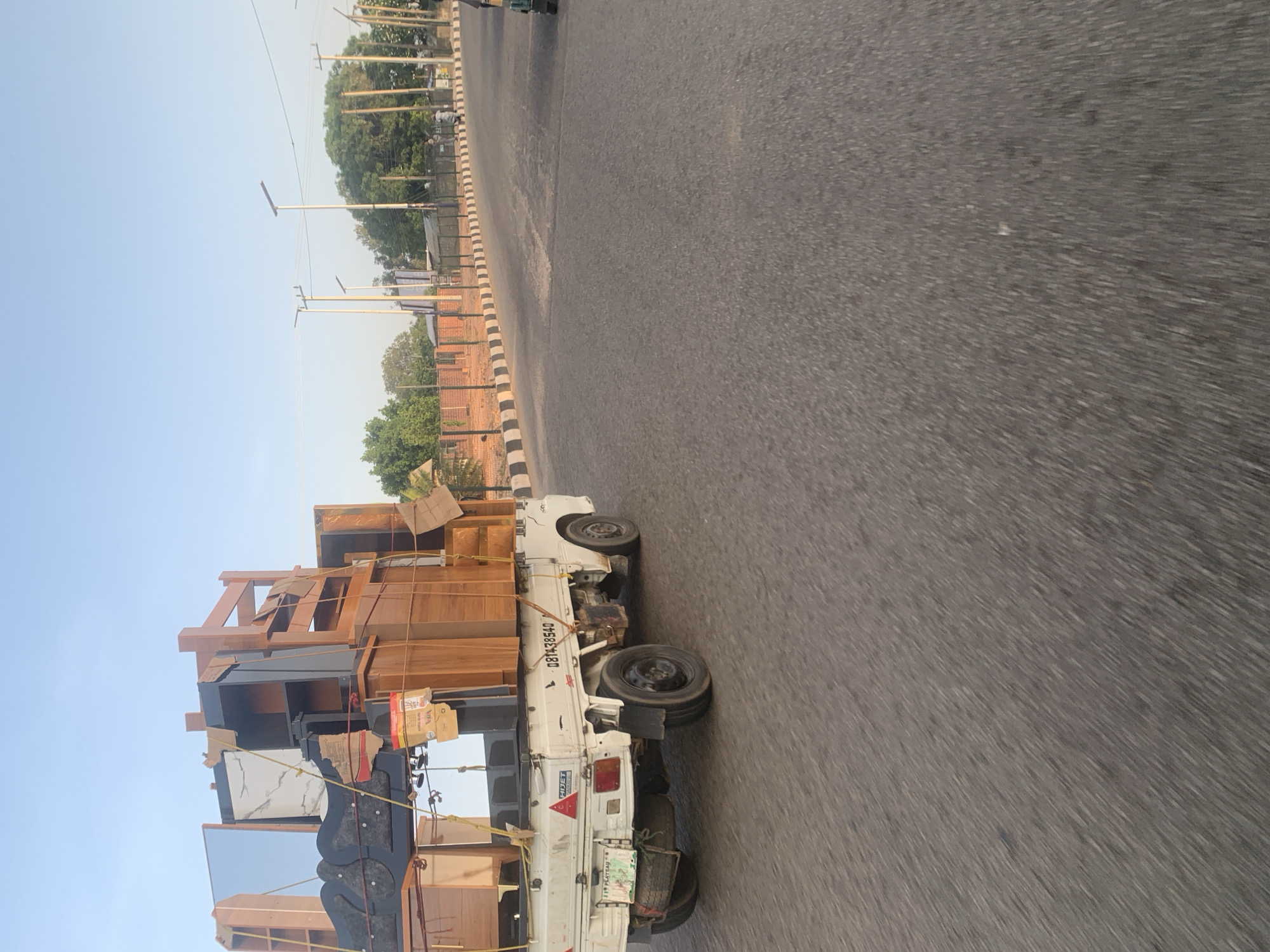 The image shows a white flatbed truck overloaded with furniture, including wooden cabinets, shelves, and what appears to be a large mirror. The furniture is secured with ropes and some cardboard. The truck has a license plate visible, reading "081438540" and "PLATEAU". It is driving on a paved road in Jos, Nigeria, with streetlights and trees lining the side of the road. A brick wall and fence are also visible. The sky is clear and blue, suggesting it is daytime. There are no people clearly visible in the immediate vicinity of the truck. The overall scene depicts the transportation of goods within the city.