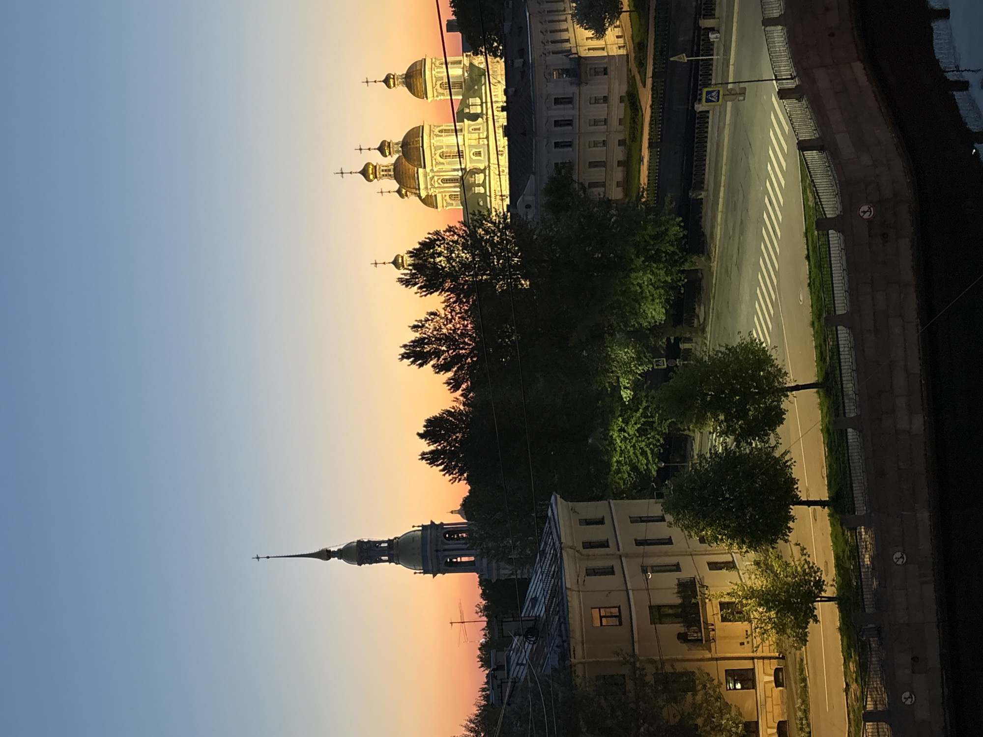 This image captures a twilight scene in Saint Petersburg, Russia. The sky transitions from a clear blue to a warm orange and pink hue near the horizon. The Smolny Cathedral, characterized by its blue and white facade and golden domes, dominates the upper section of the frame. In the lower part of the picture, the Peter and Paul Cathedral is visible, recognized by its tall, needle-like spire. A street runs between the two cathedrals, complete with pedestrian crossings and traffic signs. Buildings line the street. Trees are interspersed, and a river embankment is visible to the right.