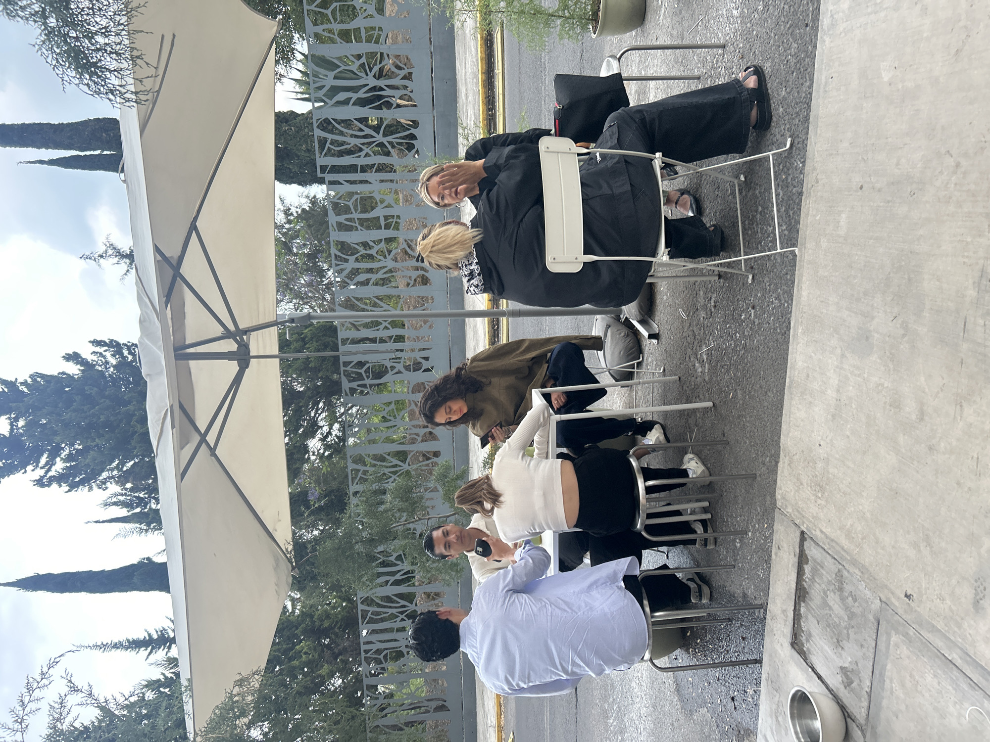 The image shows six people sitting in chairs at an outdoor café or restaurant in Puebla, Mexico. They are positioned around a table, though the table itself is not fully visible. A large umbrella provides shade for the group. The people appear to be engaged in conversation or other social interaction, possibly using their mobile phones. The environment includes trees and architectural fences. The location is outdoors and likely in an urban area of Puebla.