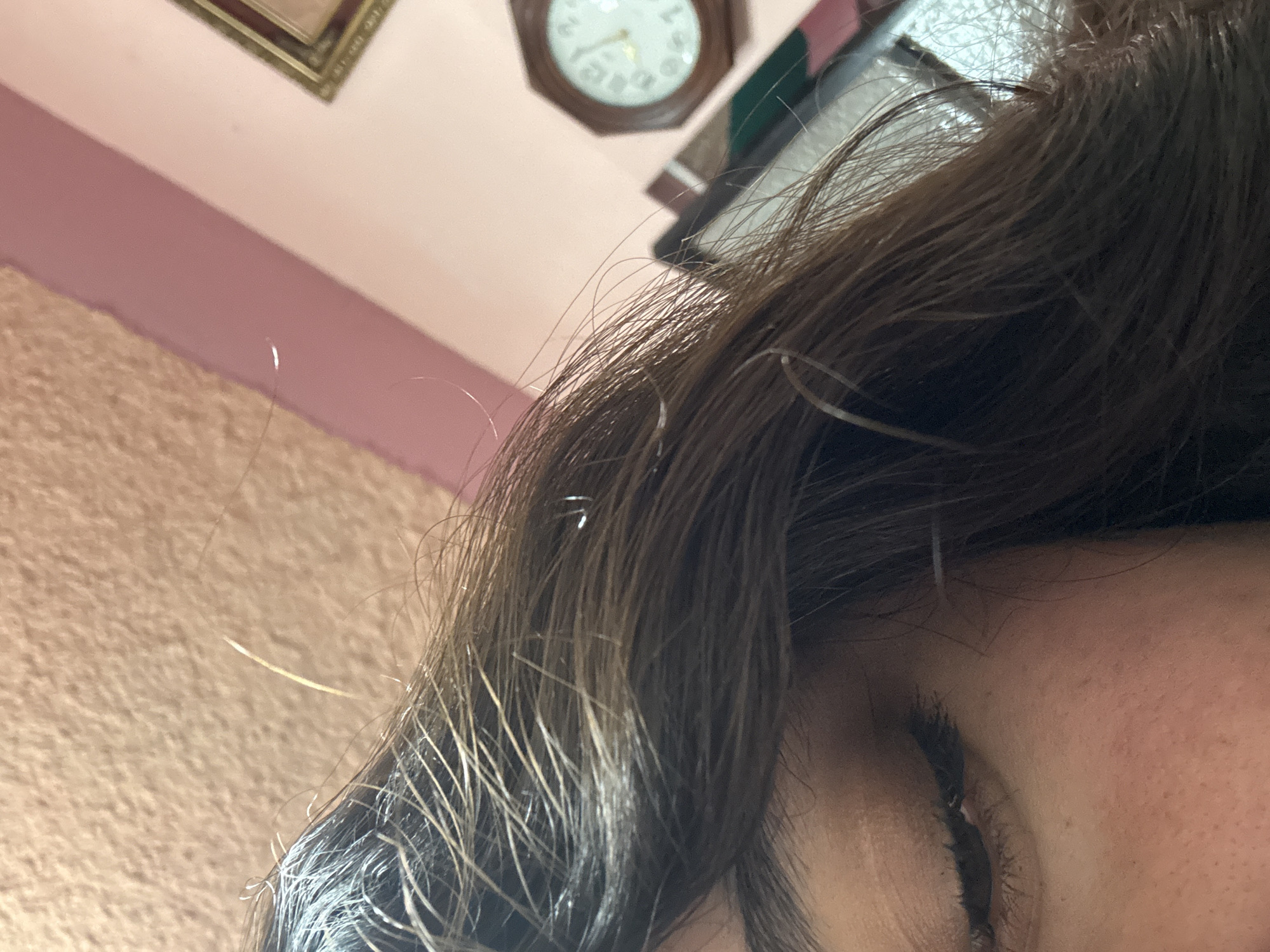The image is a close-up of a person's face, focusing on the right eye and a portion of their dark brown hair. The person's eye is partially open, revealing dark eyelashes. The hair appears to be slightly oily or wet, with individual strands visible.
In the background, the upper portion of a beige wall is visible, with a section painted light pink. A framed picture hangs on the wall. Above the hair, a round clock with roman numerals is visible. The location cannot be determined from the information provided.