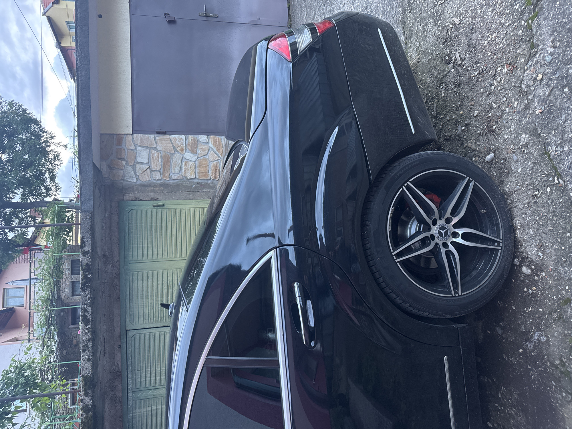 A black Mercedes-Benz S-Class is parked on a paved surface next to a building in Vratsa, Bulgaria. The building has a gray metal garage door, a stone facade, and a green wooden door. The car's rear passenger side is visible, showcasing a multi-spoke wheel. The sky overhead is overcast. The general location can be estimated with Vratsa, Bulgaria because it is the only location provided.