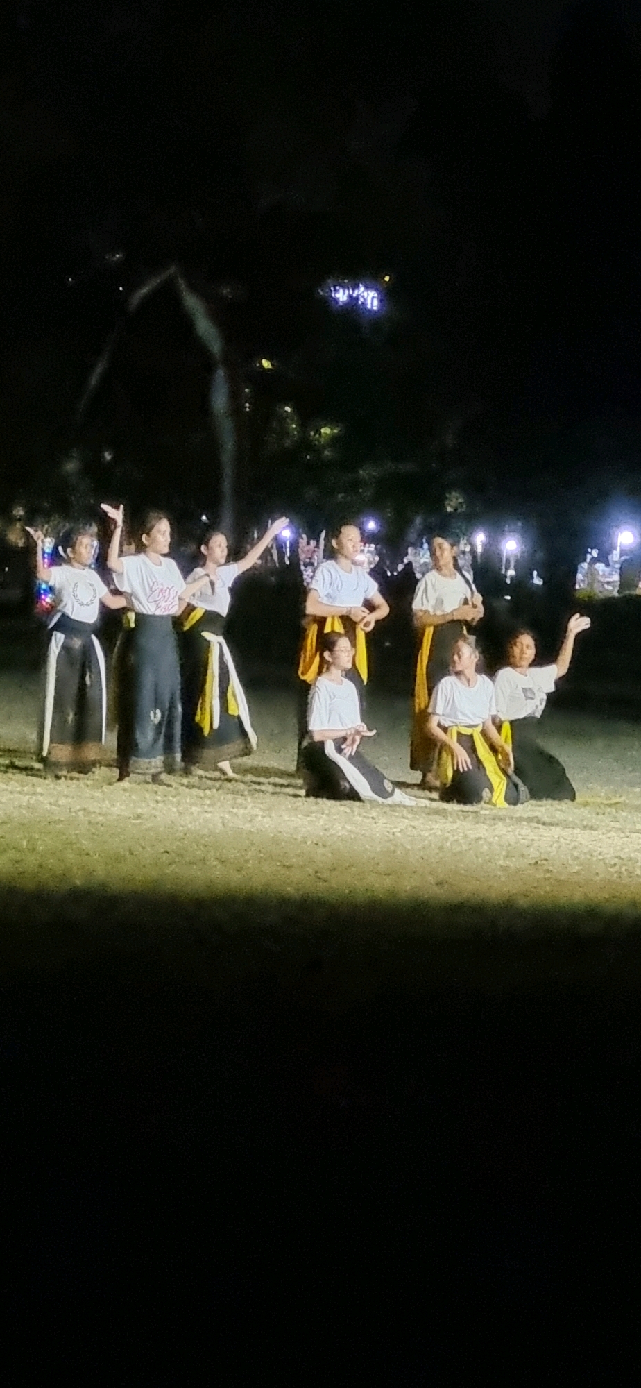 Eight young females are performing a dance routine outdoors at night in Denpasar, Indonesia. They are on a grassy field, illuminated by stage lighting and distant, blurry lights that suggest an urban environment or a festival. The dancers are wearing white t-shirts and dark, floor-length skirts with accents of black and yellow. Some are standing, while others are kneeling or in seated poses, suggesting a choreographed performance. The background is dark, with some faint illumination from what appears to be trees or structures in the distance.