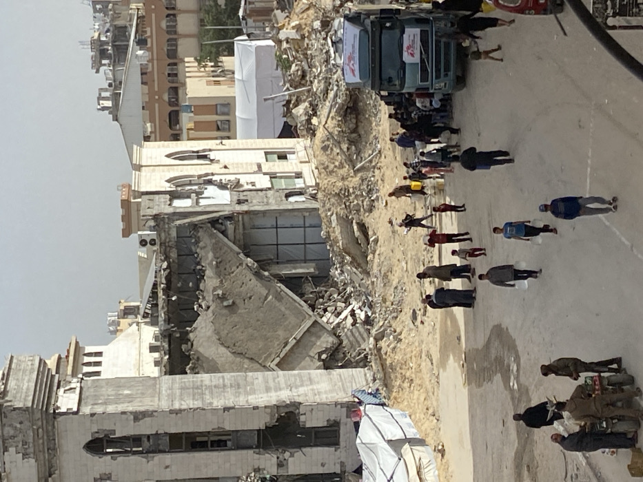 The image captures a severely damaged urban landscape in Khan Yunis, Palestinian Territory, under daylight conditions. In the background, multiple multi-story buildings exhibit extensive destruction, featuring collapsed sections, exposed structural elements, and large piles of rubble. A notable building on the left side displays a partially destroyed tower-like structure and significant facade damage. An adjacent building shows a large concrete slab, possibly a roof or floor, tilted into a substantial debris field.
In the mid-ground, a broad expanse of sandy ground covered with rubble separates the damaged structures from the foreground activity. A large white tent is visible in the background, amidst the rubble, and a smaller white tent with blue tarpaulin elements is present to the left, closer to the damaged building.
The foreground features a light-colored dirt road occupied by approximately 30-40 civilians, comprising adults and children, dressed in casual clothing. On the right side of the road, a light blue/grey Volvo semi-truck, adorned with "Médecins Sans Frontières" (MSF) banners on its roof and side, is parked. A crowd of individuals, including many children, is actively gathered around the truck, engaging in what appears to be aid distribution or collection. Many individuals are seen carrying yellow jerry cans, clear plastic bottles, and white plastic bags, suggesting the collection of water or other supplies.
Several people walk along the road, some individually and others in small groups, with some carrying yellow jerry cans or plastic containers on their heads or shoulders. In the lower-left foreground, three men are gathered around a three-wheeled motorized cart loaded with items, interacting with its contents.